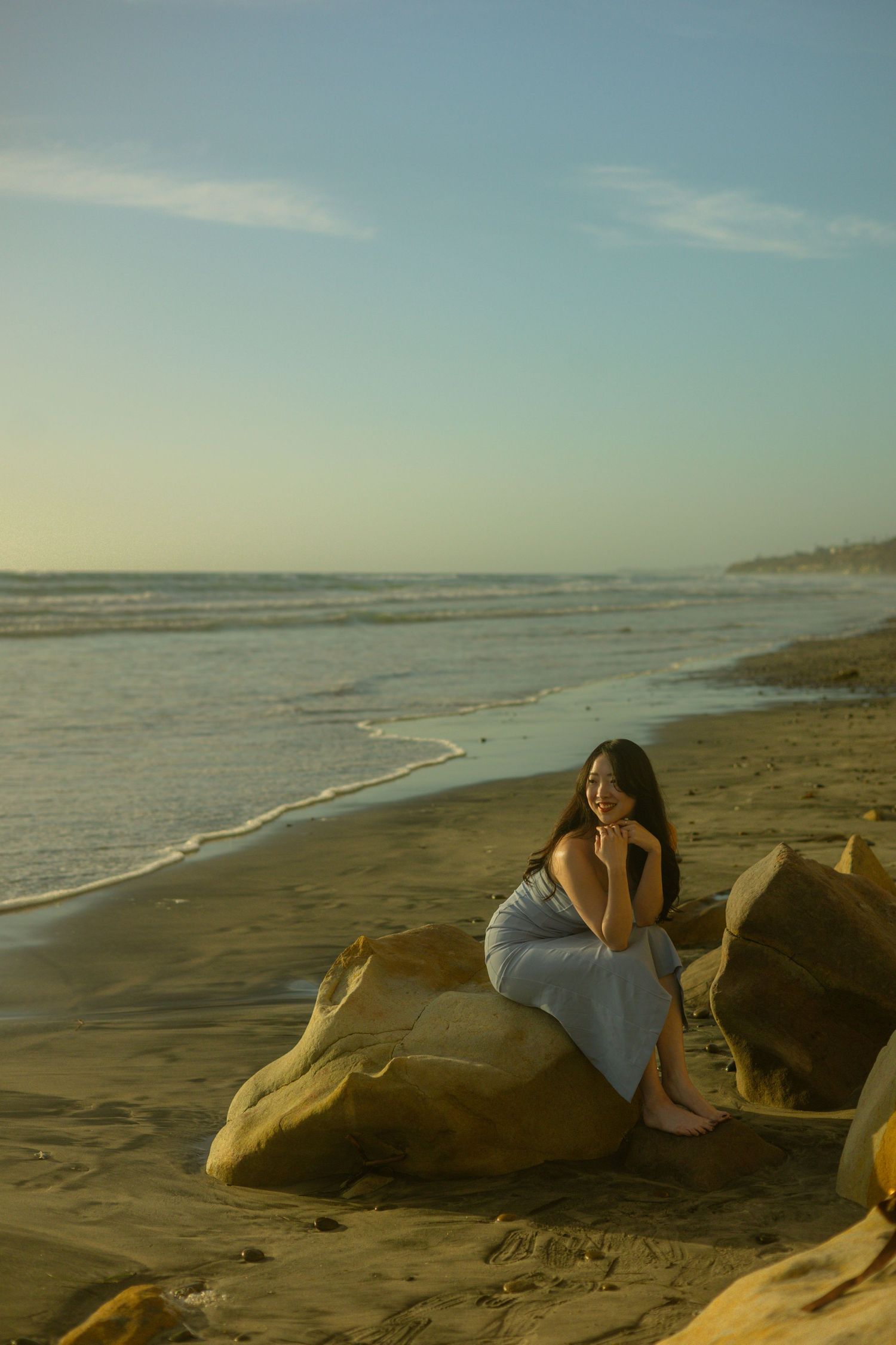 Coastal scene at dusk with figure in dress sitting on large rocks by the ocean.
