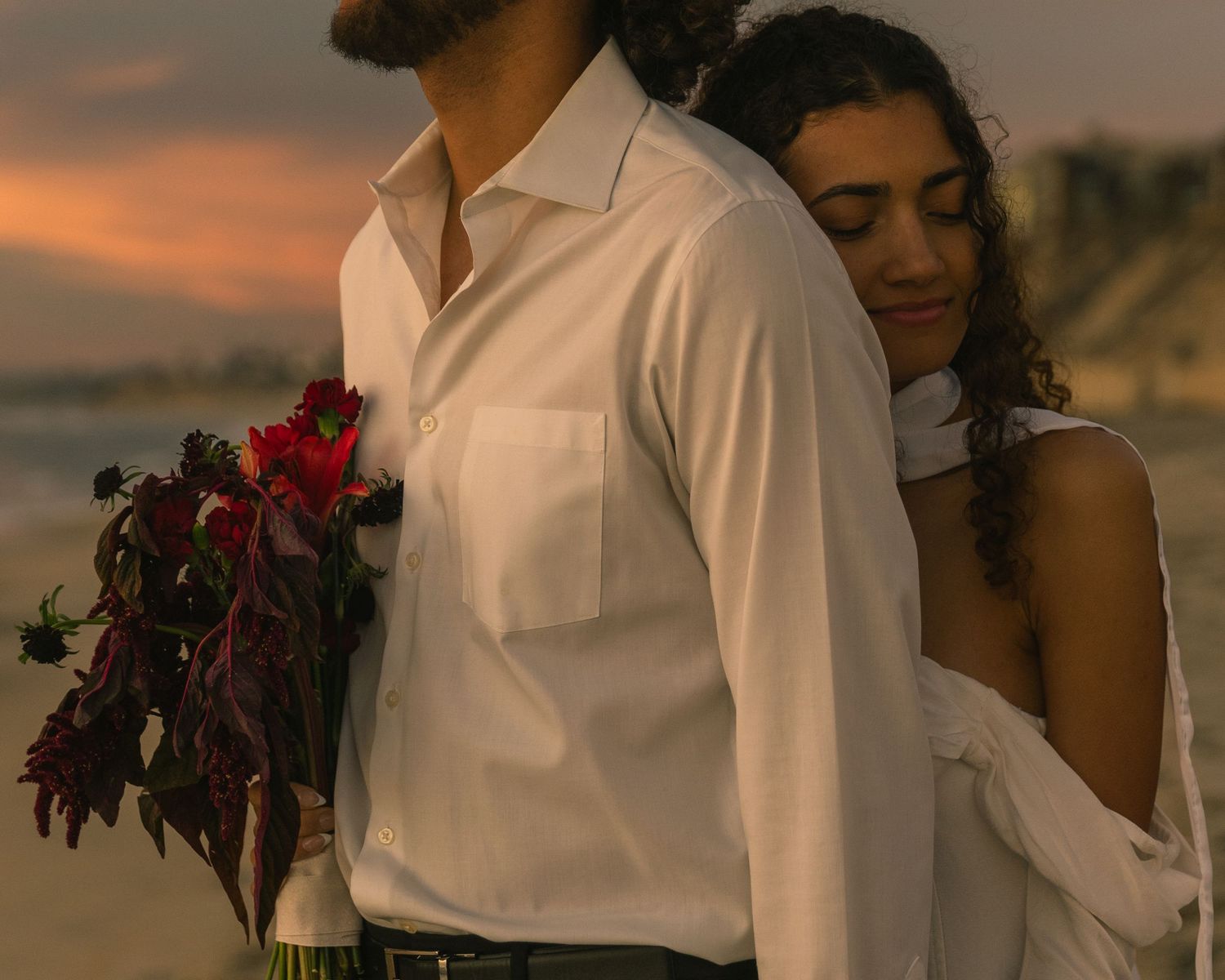 A couple in white clothing embraces during a romantic beach sunset photo session.