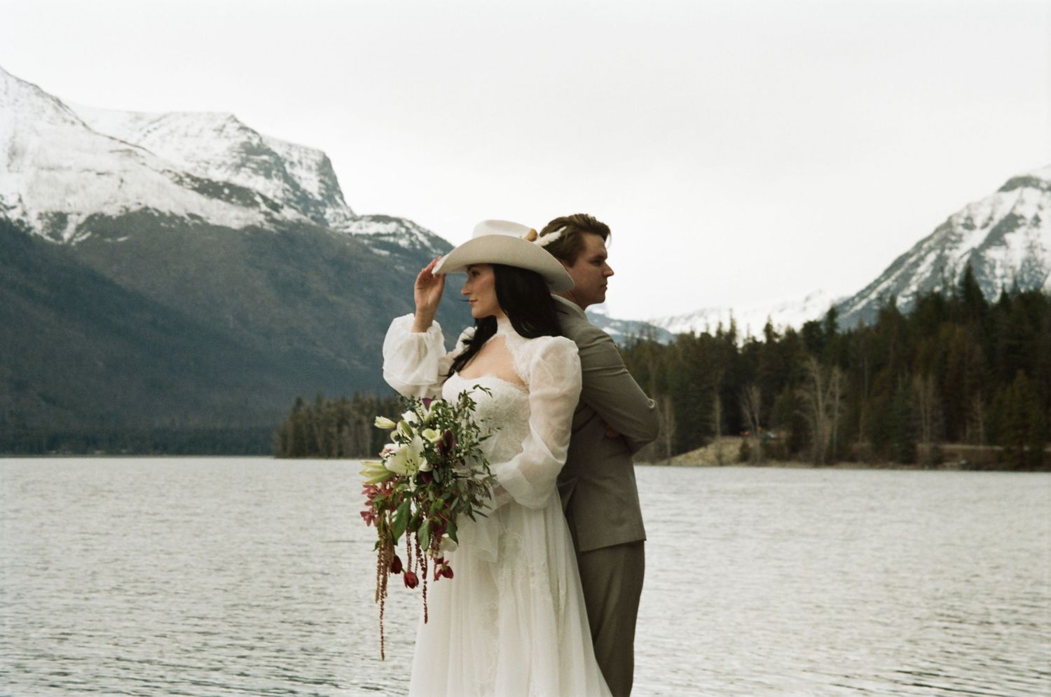 Bride and groom embracing by a mountain lake, bride holding bouquet and wearing wide-brim hat, snow-capped peaks behind.