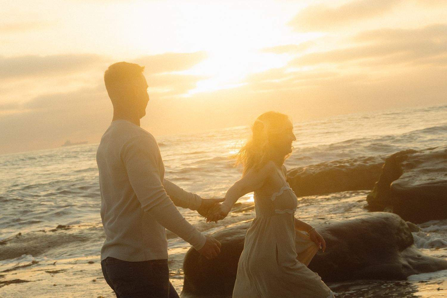 Two silhouetted figures sit on a rocky beach at sunset while ocean waves crash against the shore in the background.