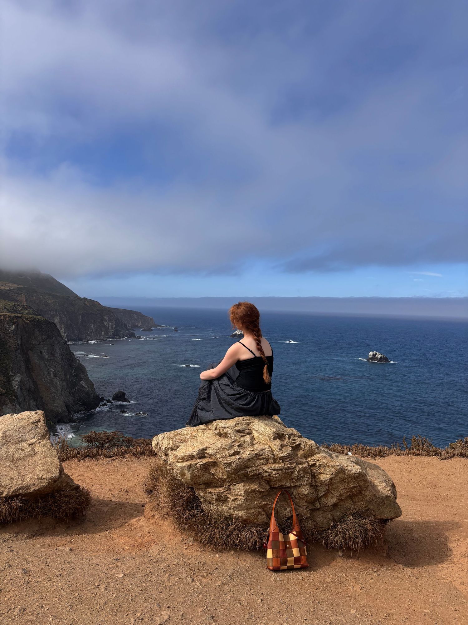 Woman in black dress sitting on a rock overlooking the dramatic Big Sur coastline with misty cliffs and blue ocean.