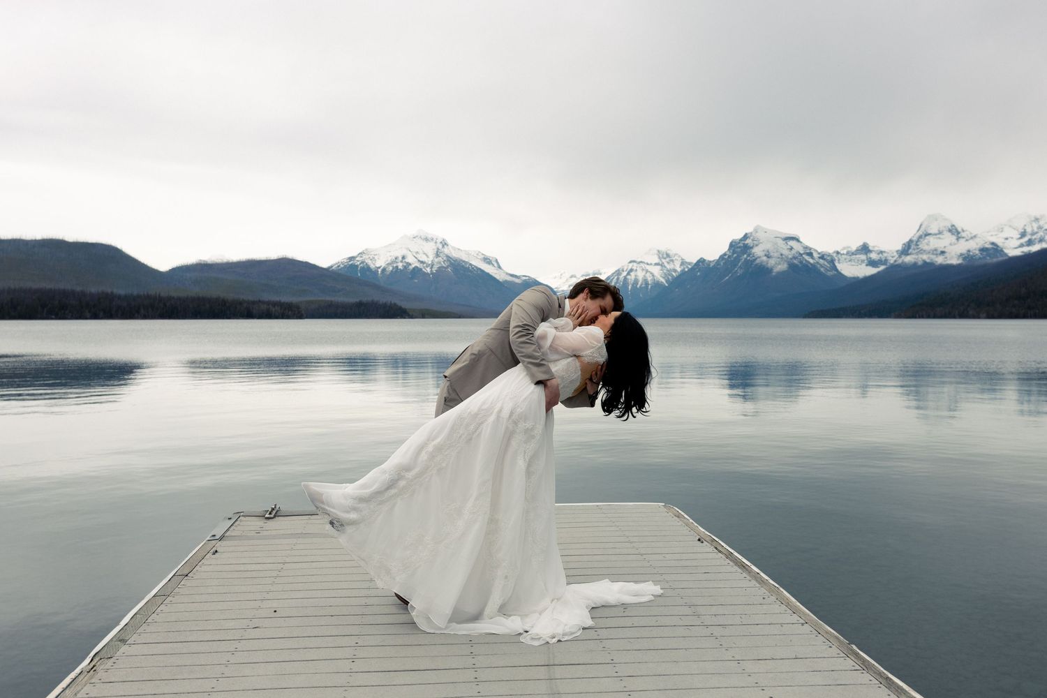 A couple in wedding attire embraces on a wooden dock overlooking a misty mountain lake with snow-capped peaks.