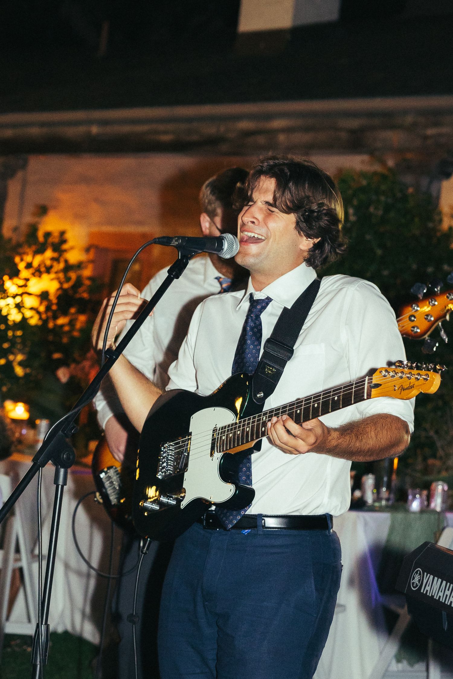 A man in a white shirt and tie plays electric guitar and sings into a microphone at an outdoor evening event.