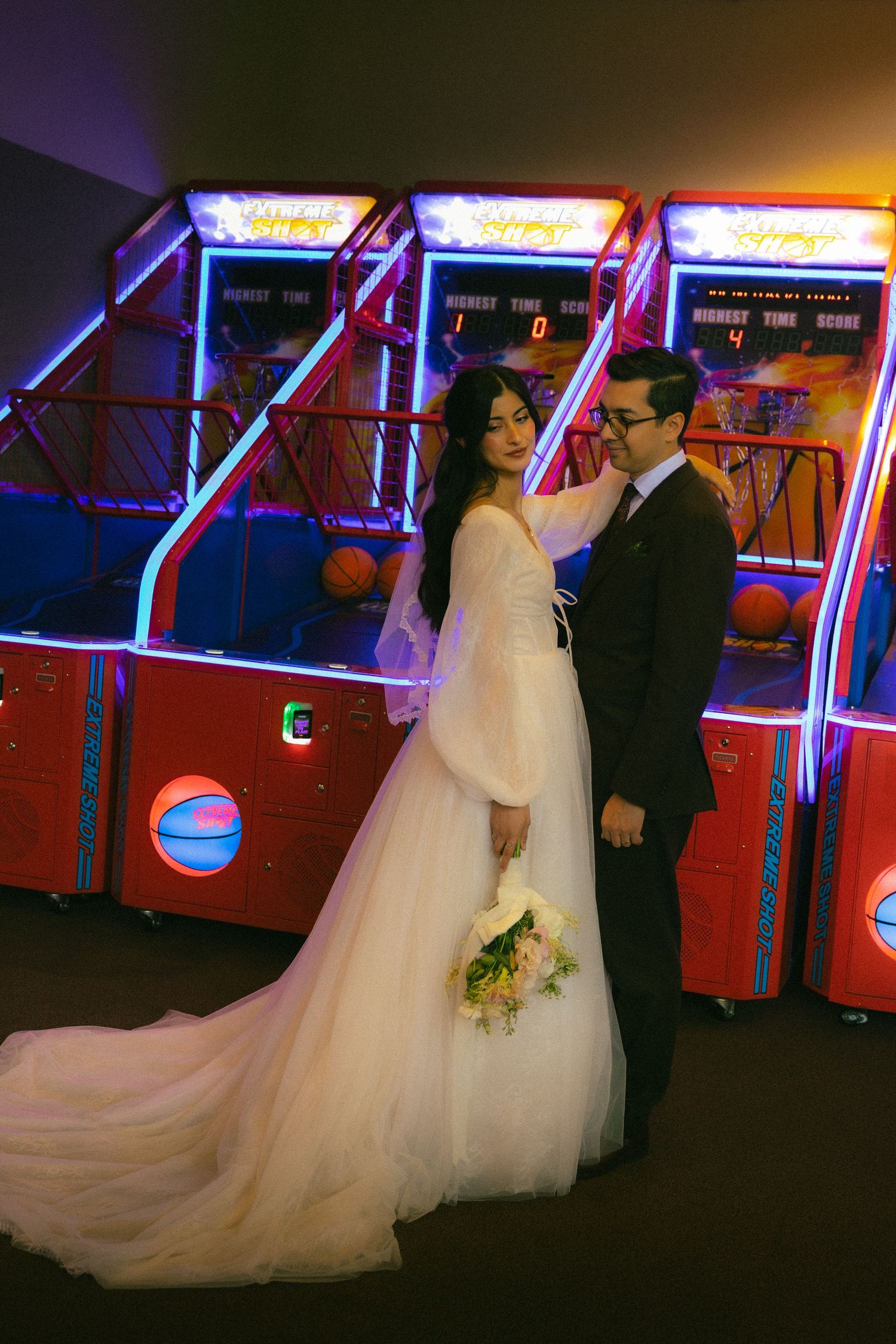 A couple in wedding attire poses in front of illuminated arcade basketball games with neon lighting.