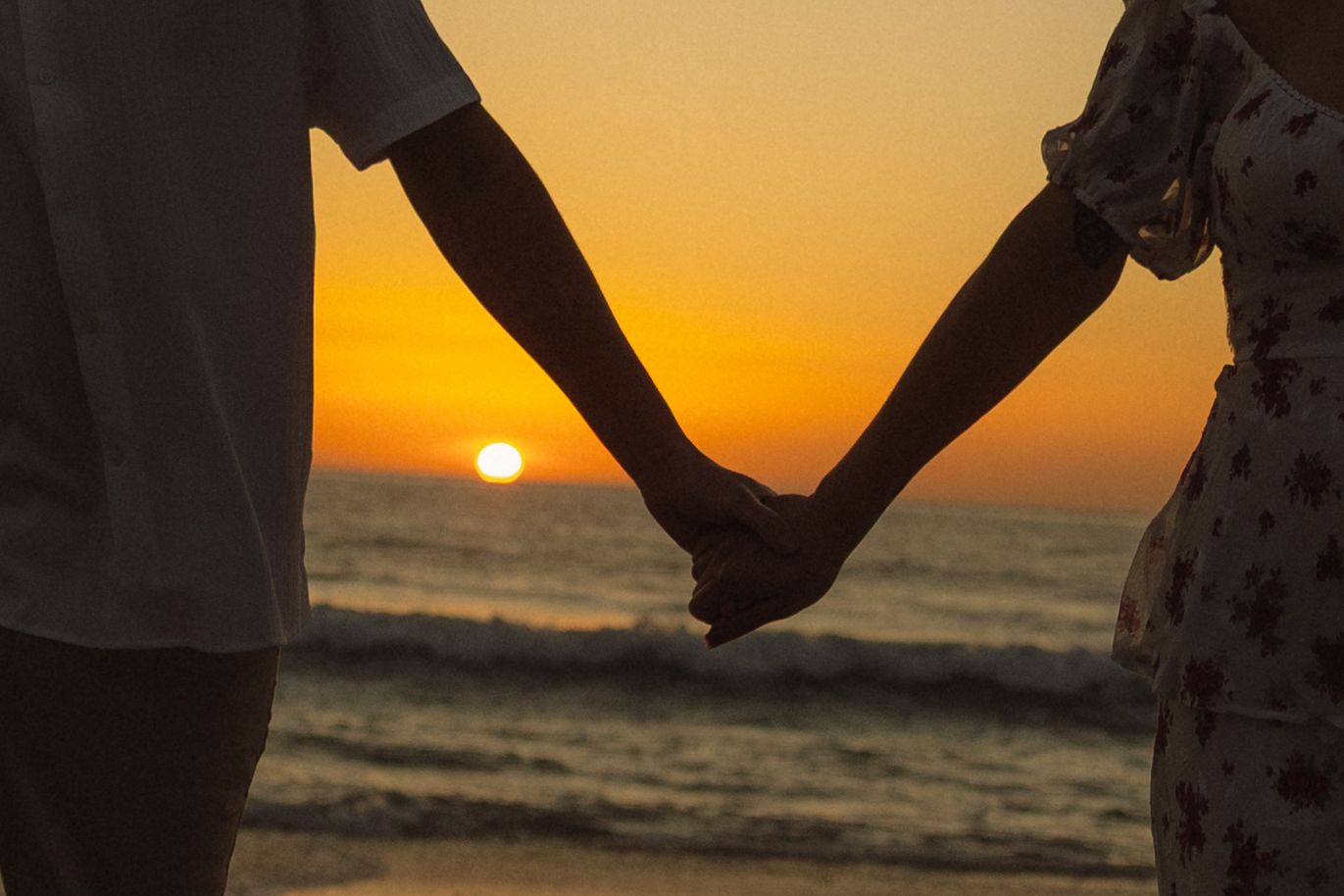 A silhouetted couple holds hands while walking on the beach at sunset with orange sky reflecting on the ocean.