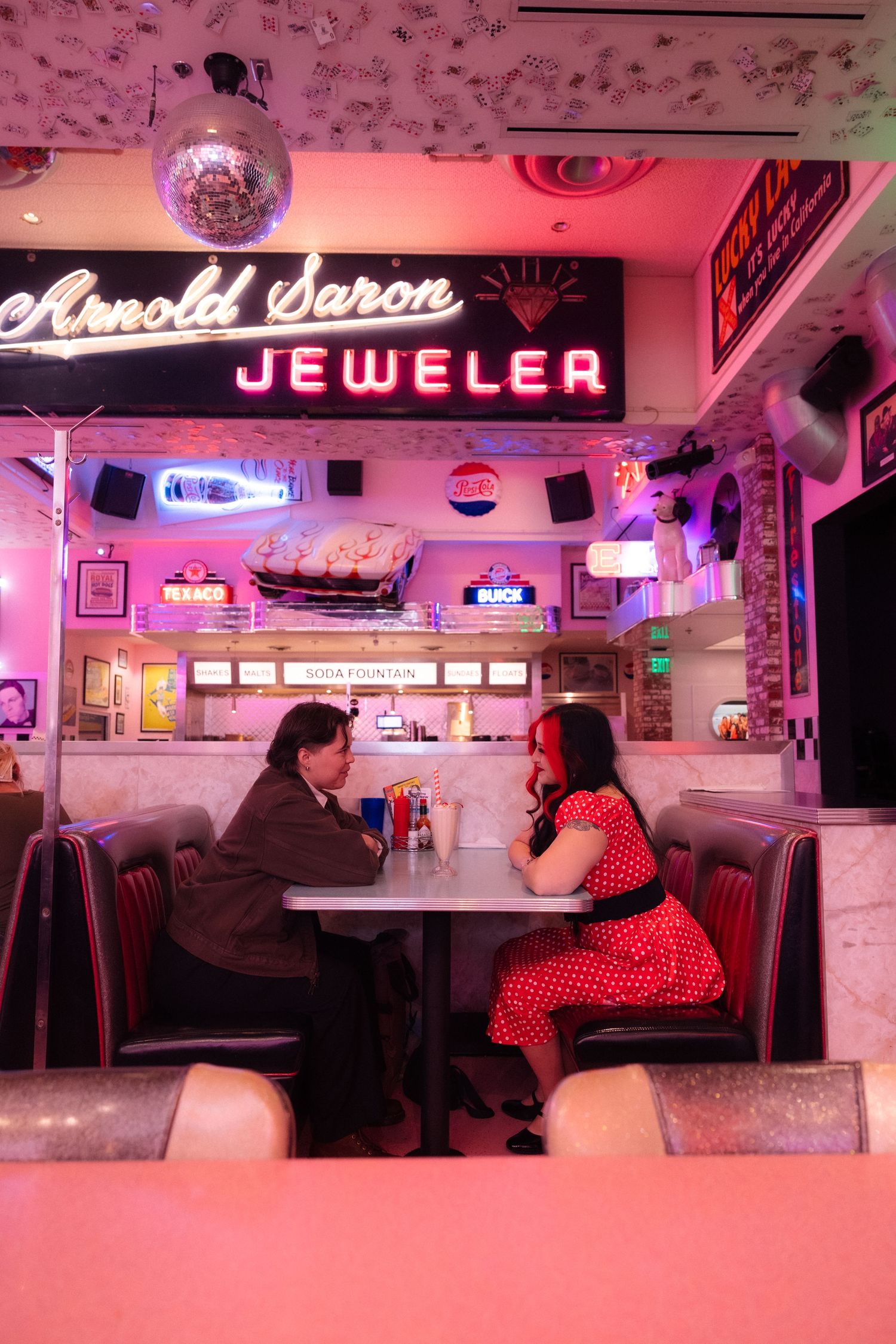 Couple dining at a retro 1950s diner with neon signs, pink lighting, and classic booth seating.