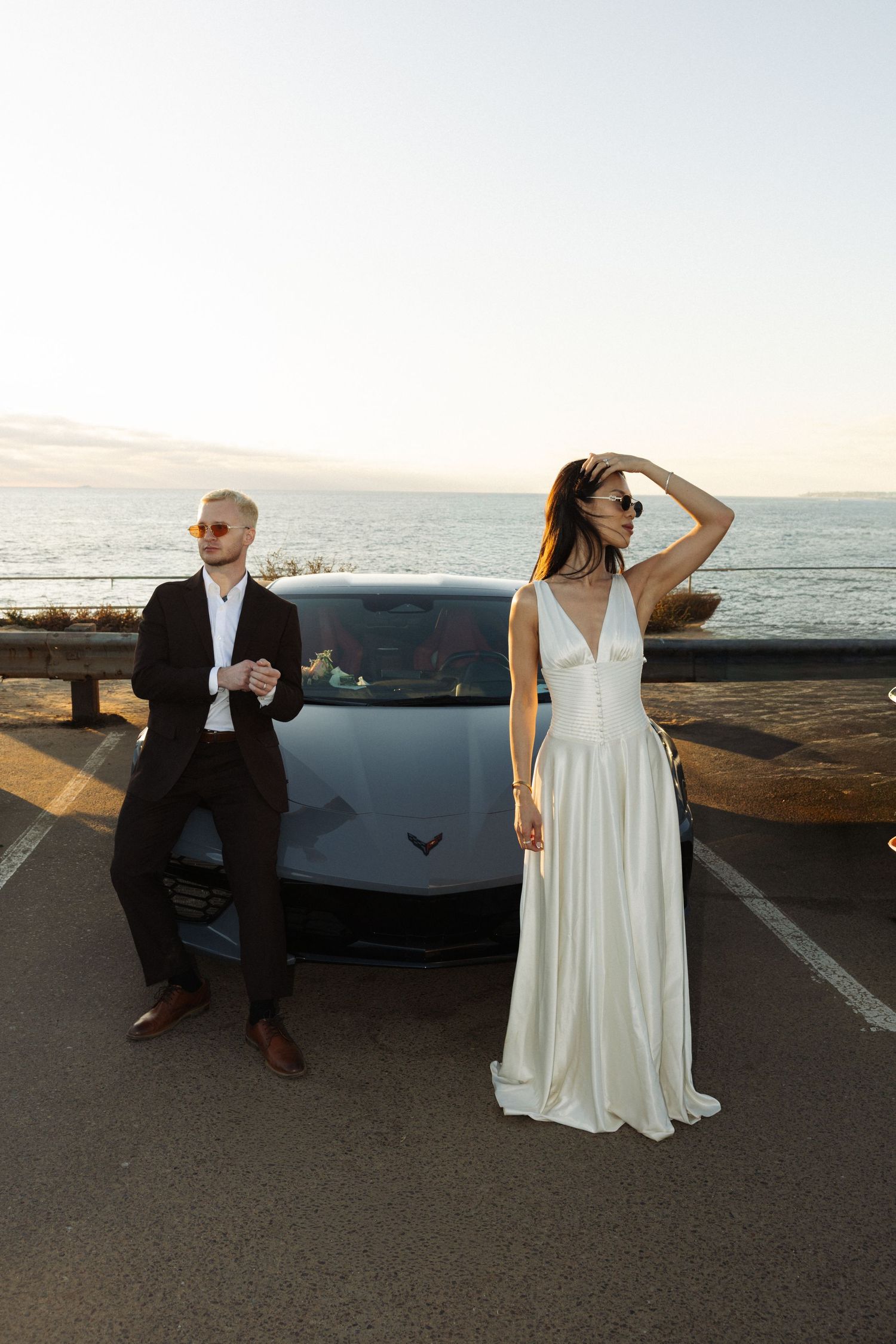 Elegant wedding couple posing by luxury sports car at sunset overlooking ocean, bride in white gown, groom in black suit.