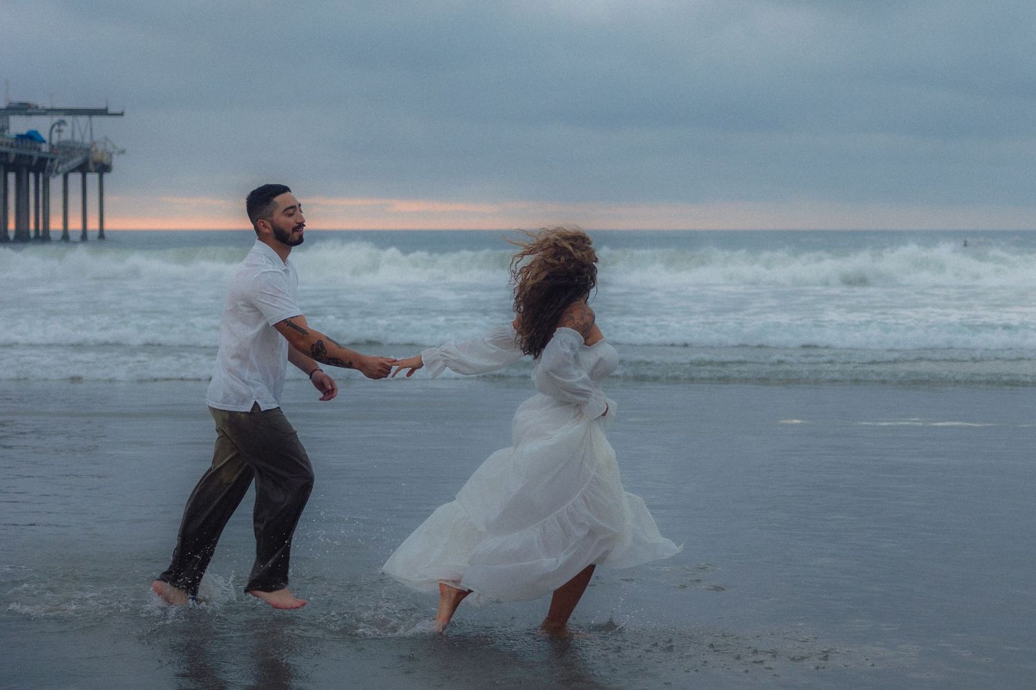 Couple in wedding attire playfully running and splashing along the beach at sunset.