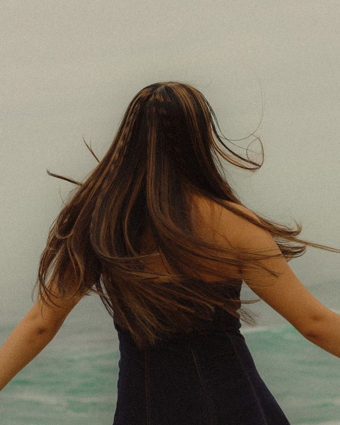 Long brown hair flowing in the wind against turquoise water backdrop, wearing a black dress in artistic motion shot.