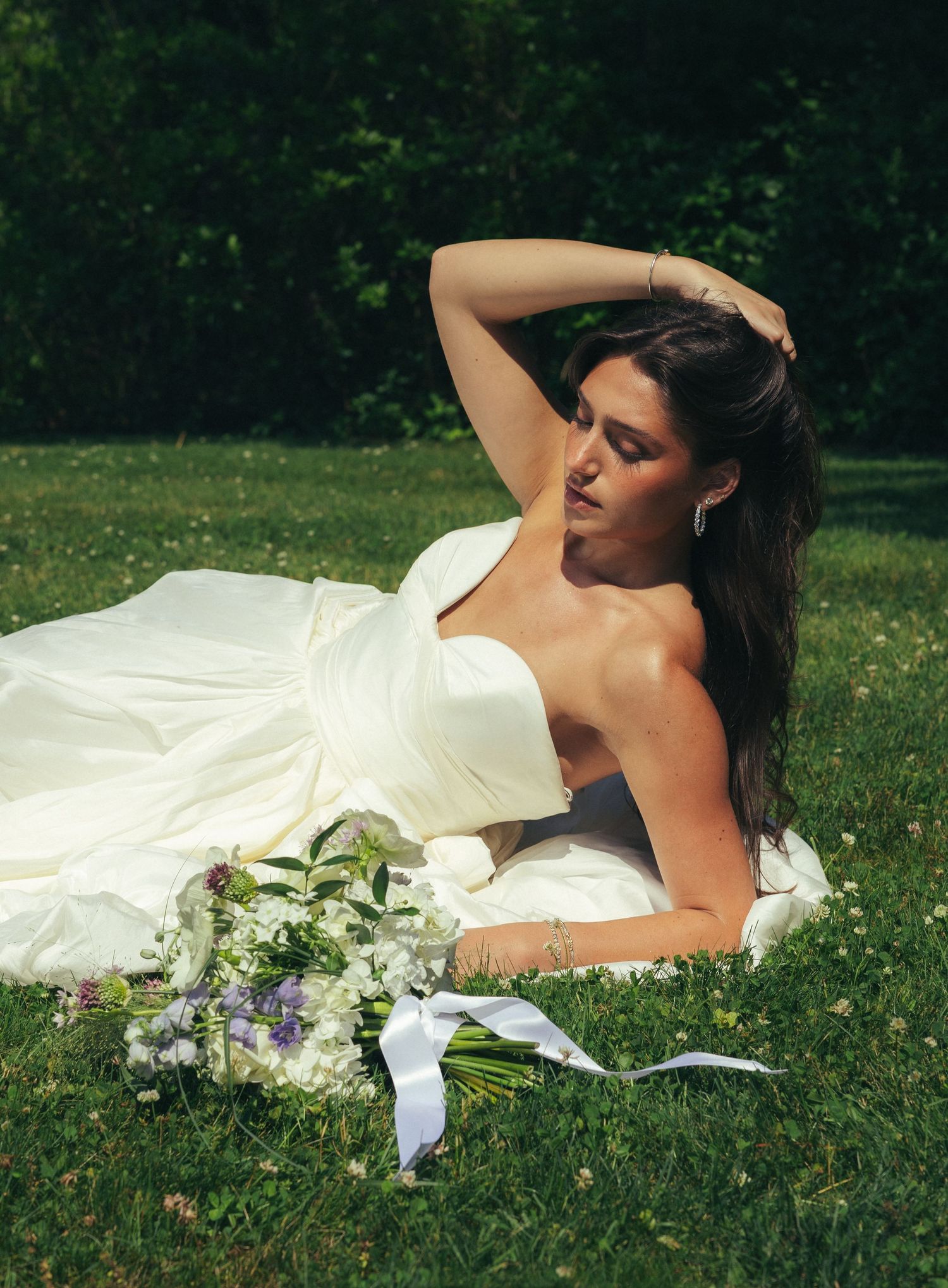 Bride in strapless ivory gown reclines on grass holding bouquet of white and purple flowers in sunny outdoor setting.