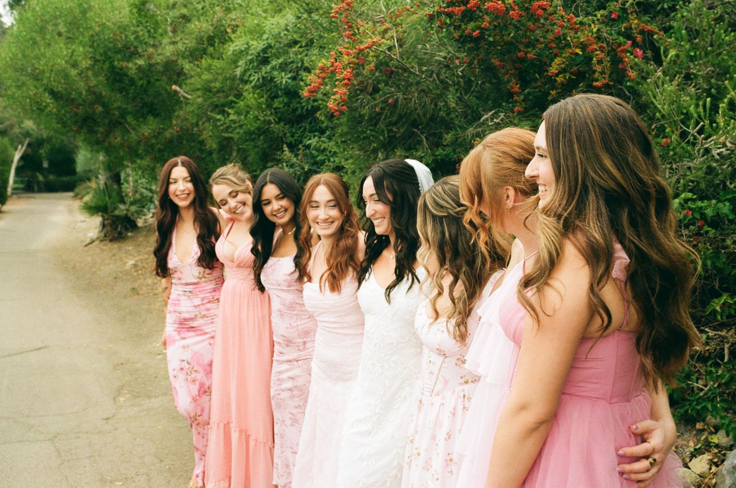 Bride and bridesmaids in pink dresses laughing together outdoors on a tree-lined path at a wedding.
