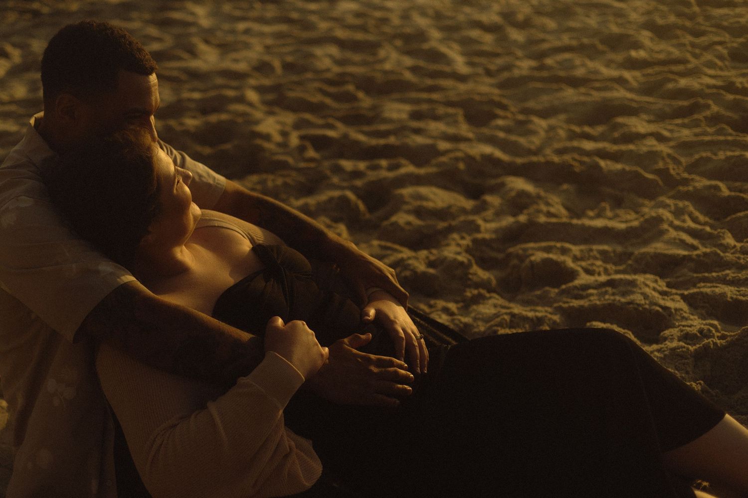 A couple embraces on sandy beach during golden hour, cast in warm sunset light and shadows.