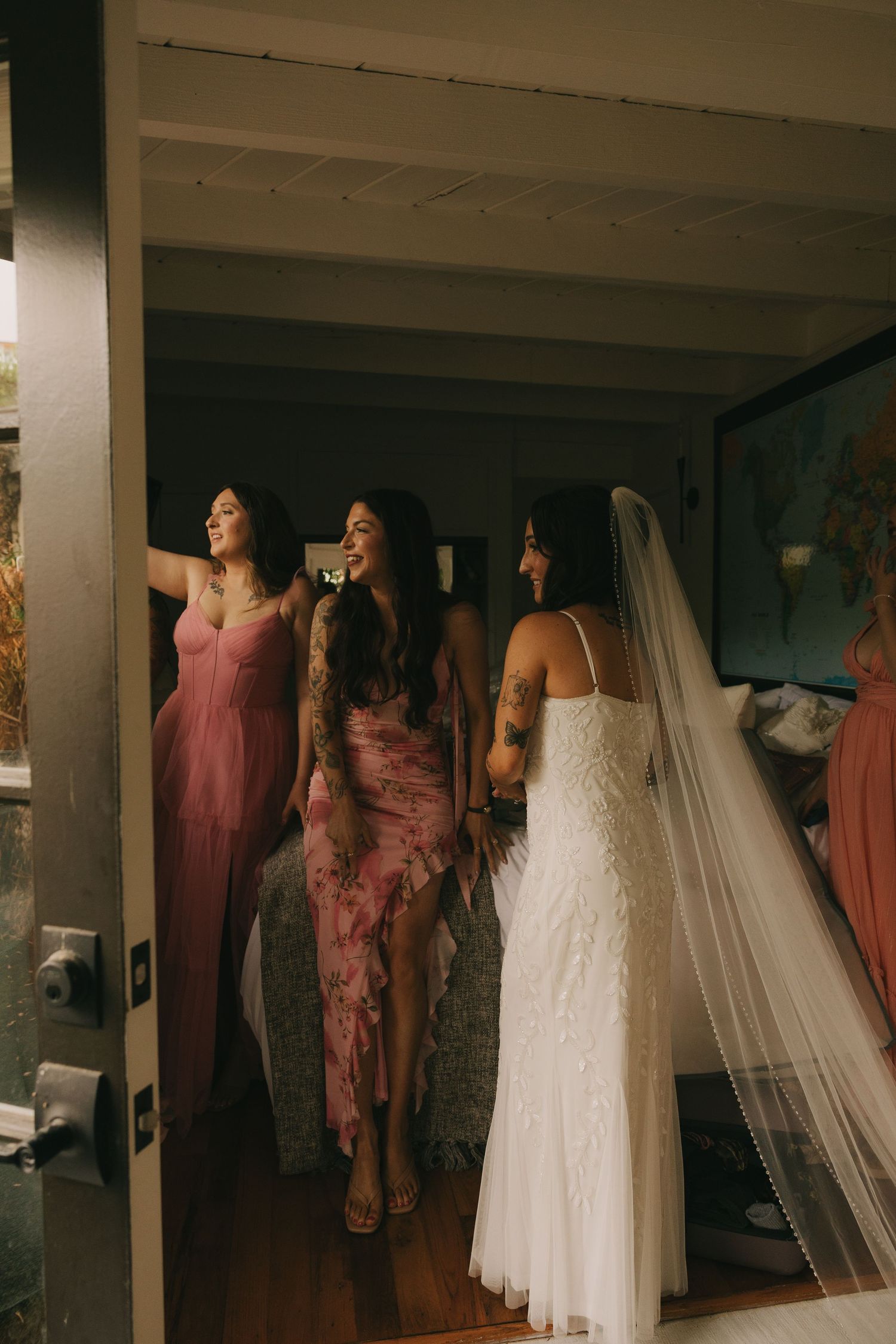 Bride in white lace gown and veil laughing with bridesmaids in pink dresses during wedding preparations.
