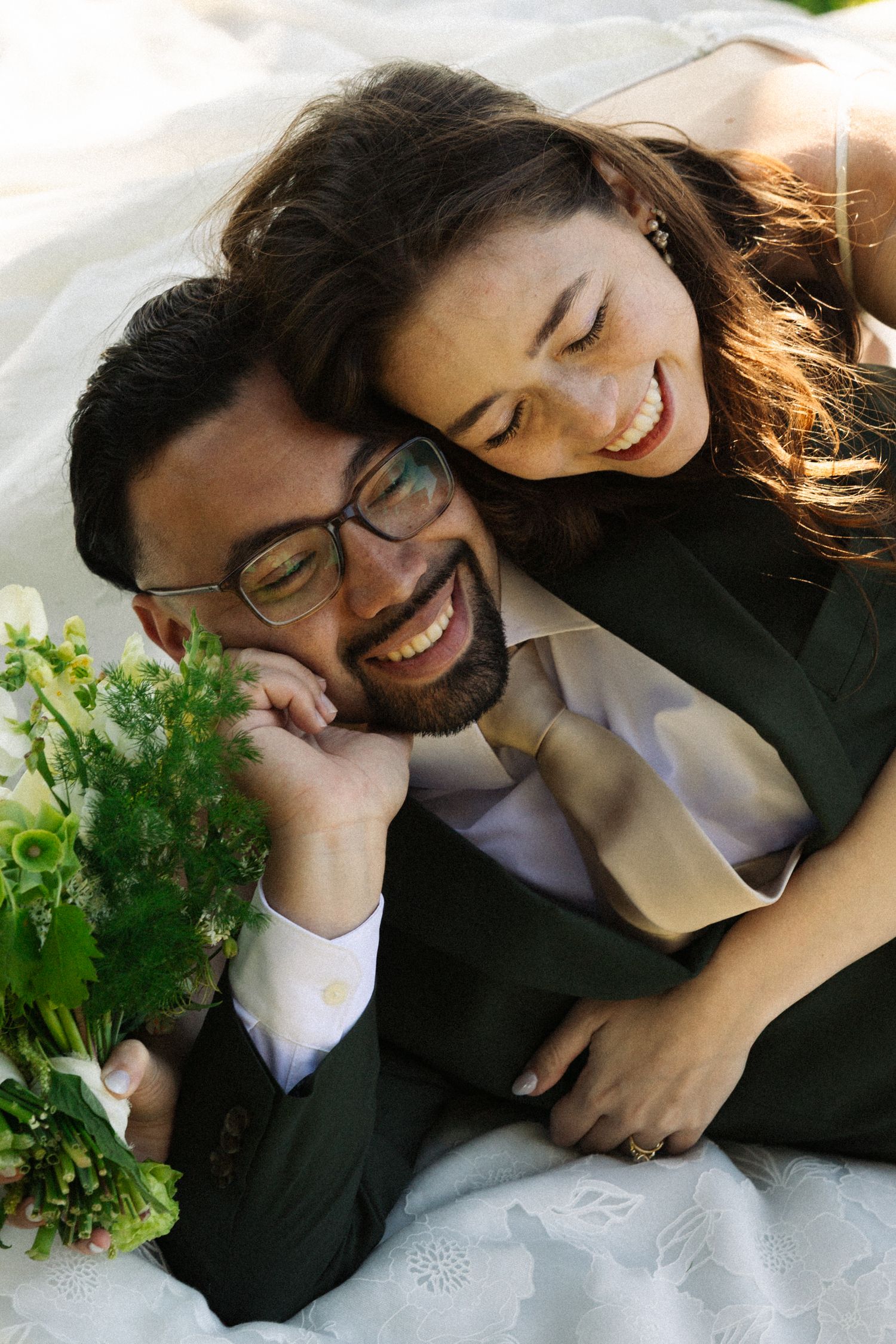 Happy couple in wedding attire laughing together outdoors, groom in dark suit with glasses holding bridal bouquet.