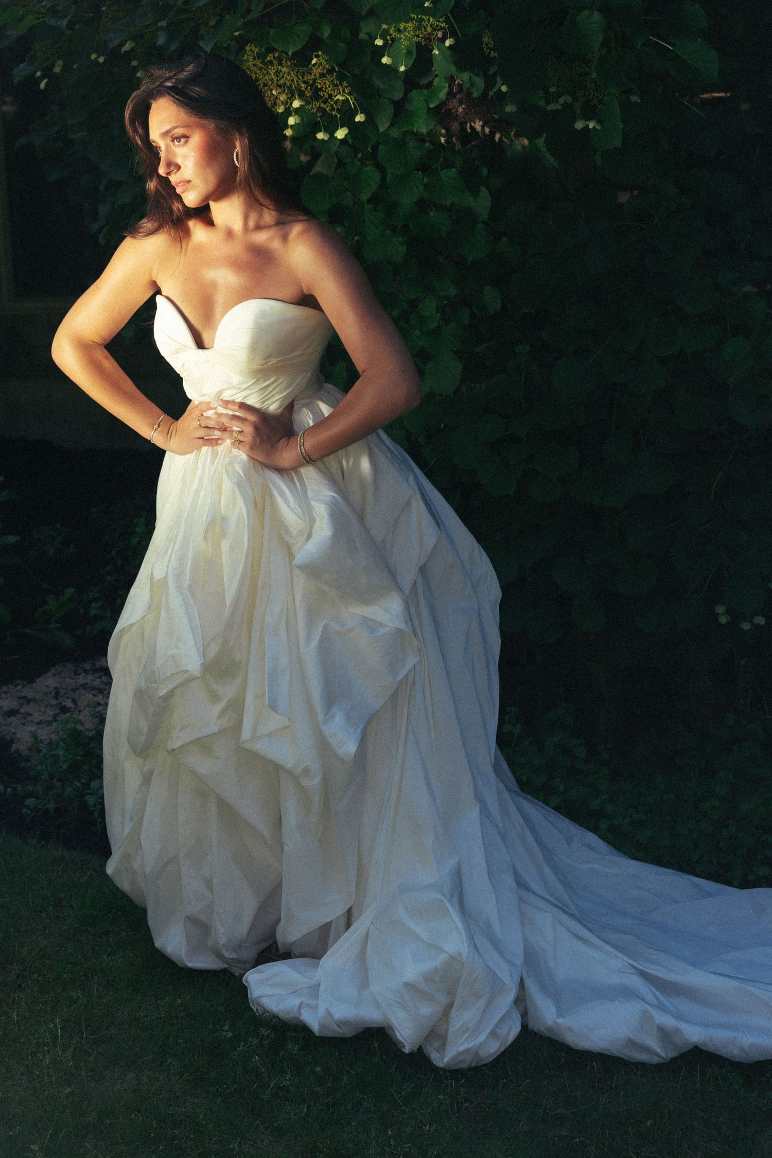 Bride in strapless ruffled white ball gown with train poses in dramatic garden lighting.