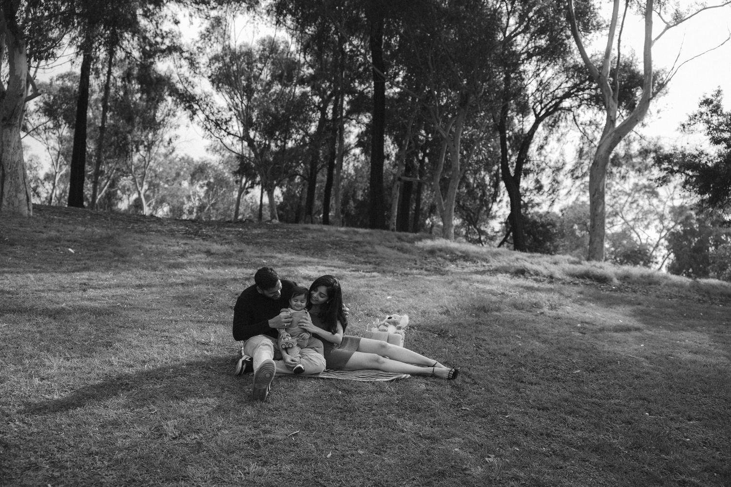 A couple sits together on a grassy hillside surrounded by trees in a black and white photograph.