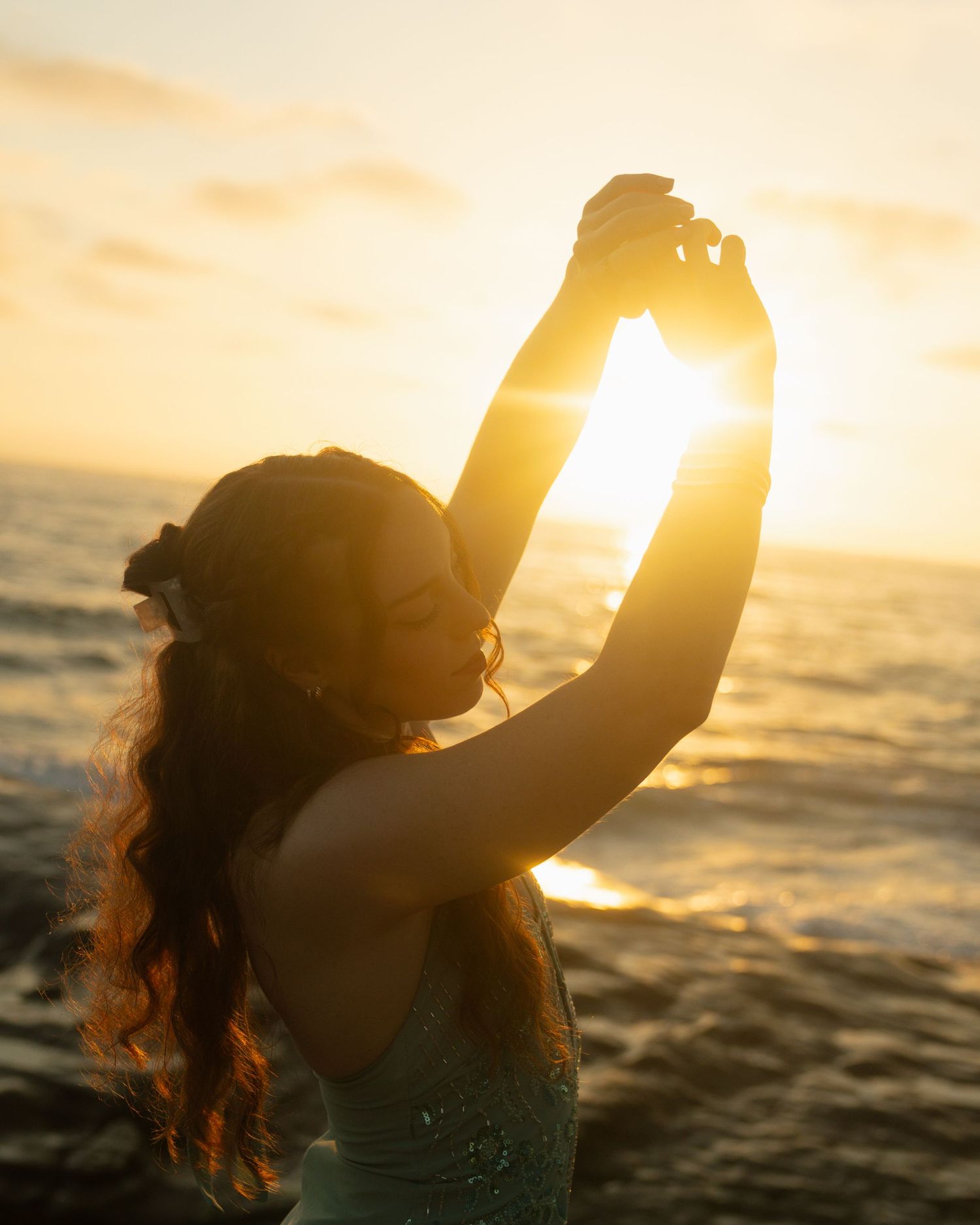 Silhouette of a person raising their arms at sunset by the ocean with golden light streaming through.