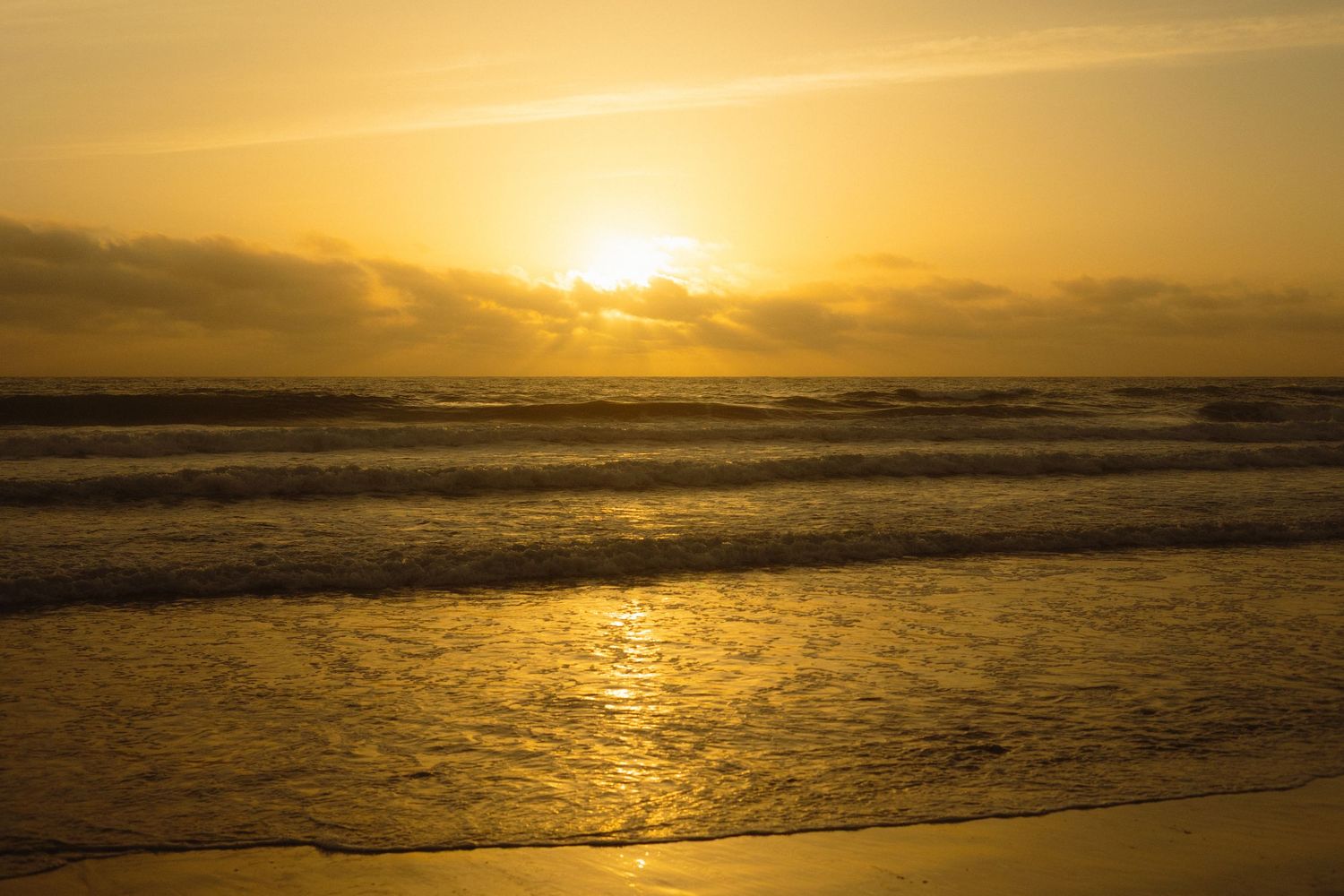 Golden sunset over ocean waves creates dramatic silhouette and reflections on wet sand at beach.
