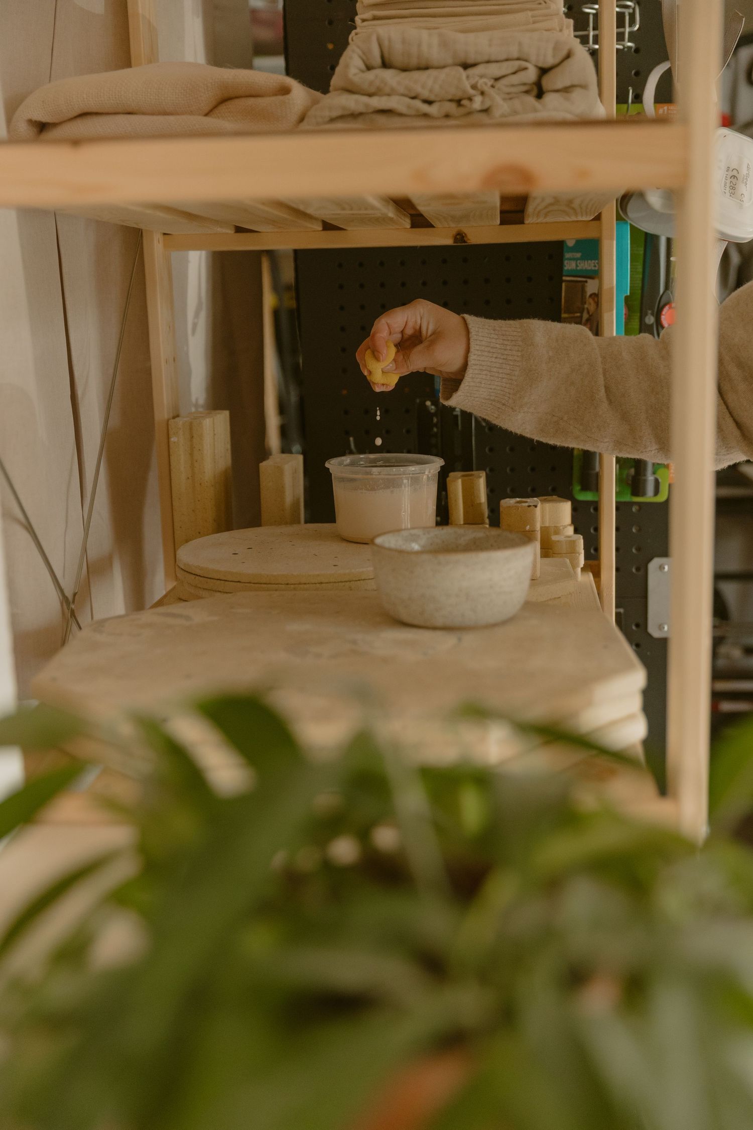 Clay artist adding water to pottery supplies on a wooden shelf with green plants in foreground.