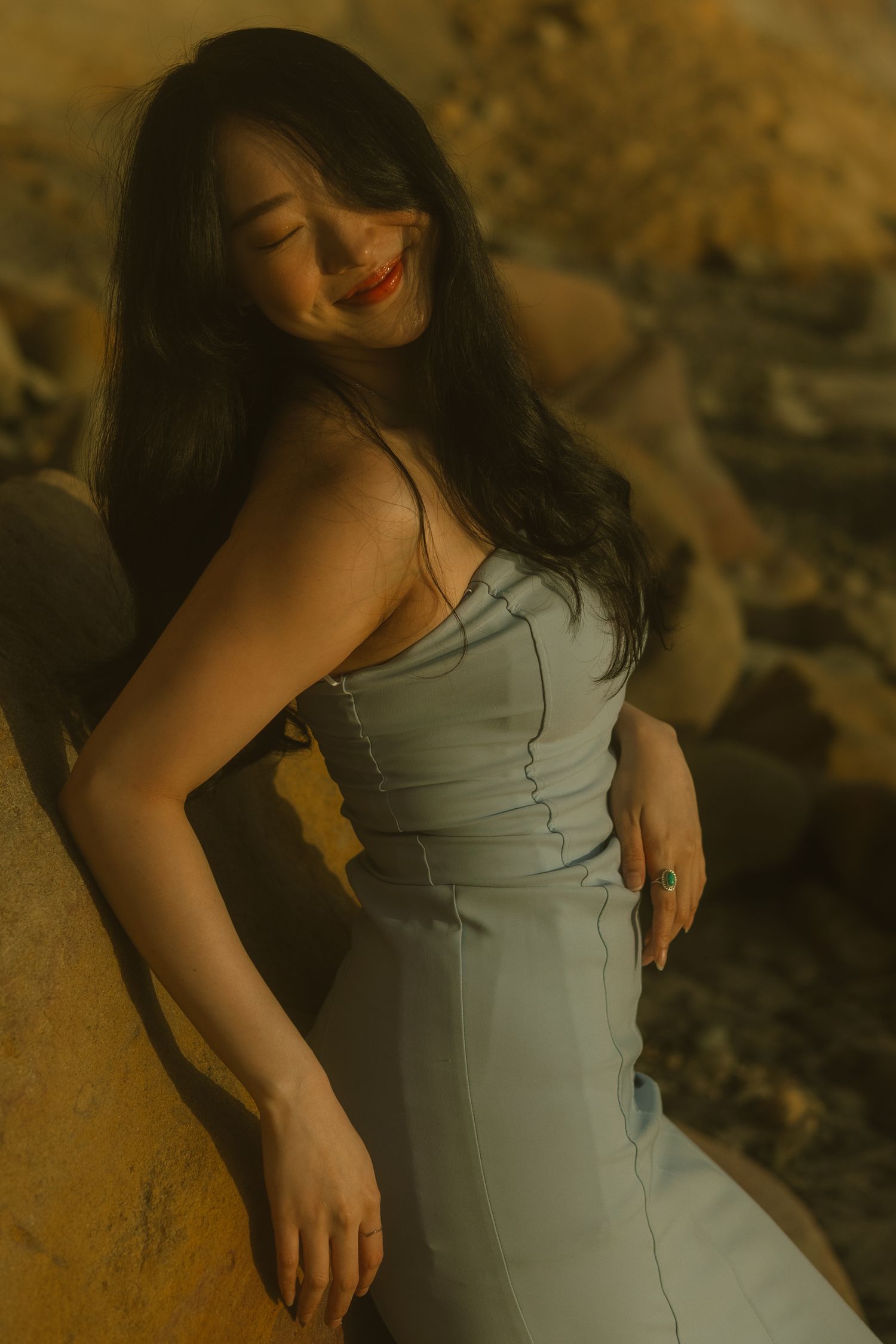 A woman in a light gray dress poses outdoors during golden hour with warm sunlit tones in the background.
