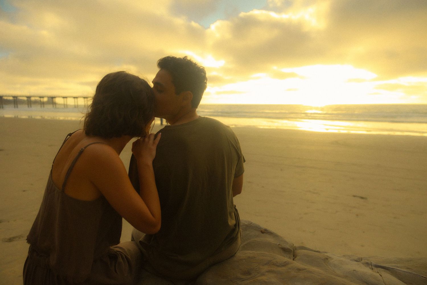 A couple shares a romantic moment on a beach at sunset with golden light illuminating the sandy shoreline.