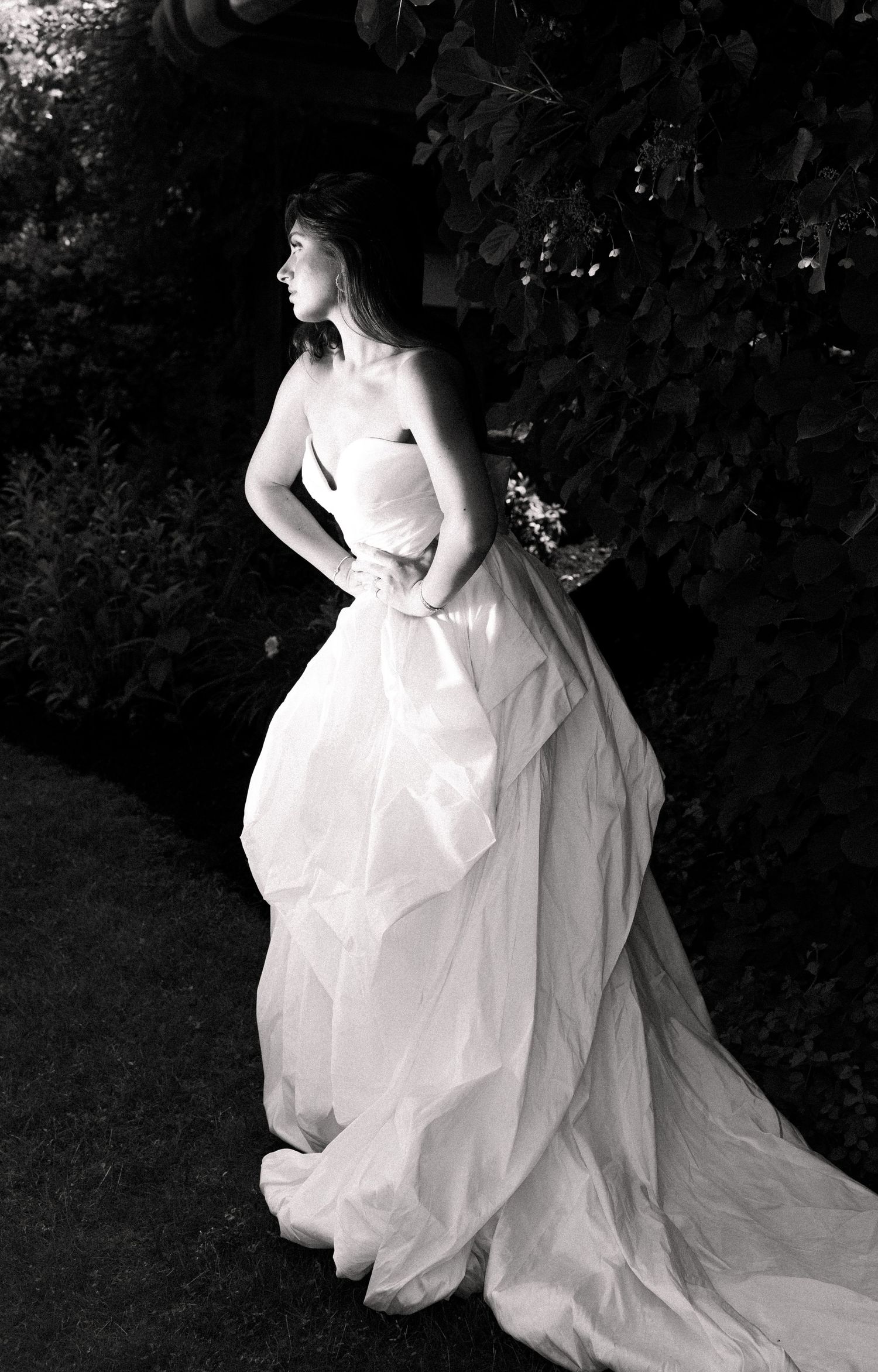 Elegant bride in voluminous strapless white ball gown stands in shadowy garden in black and white.