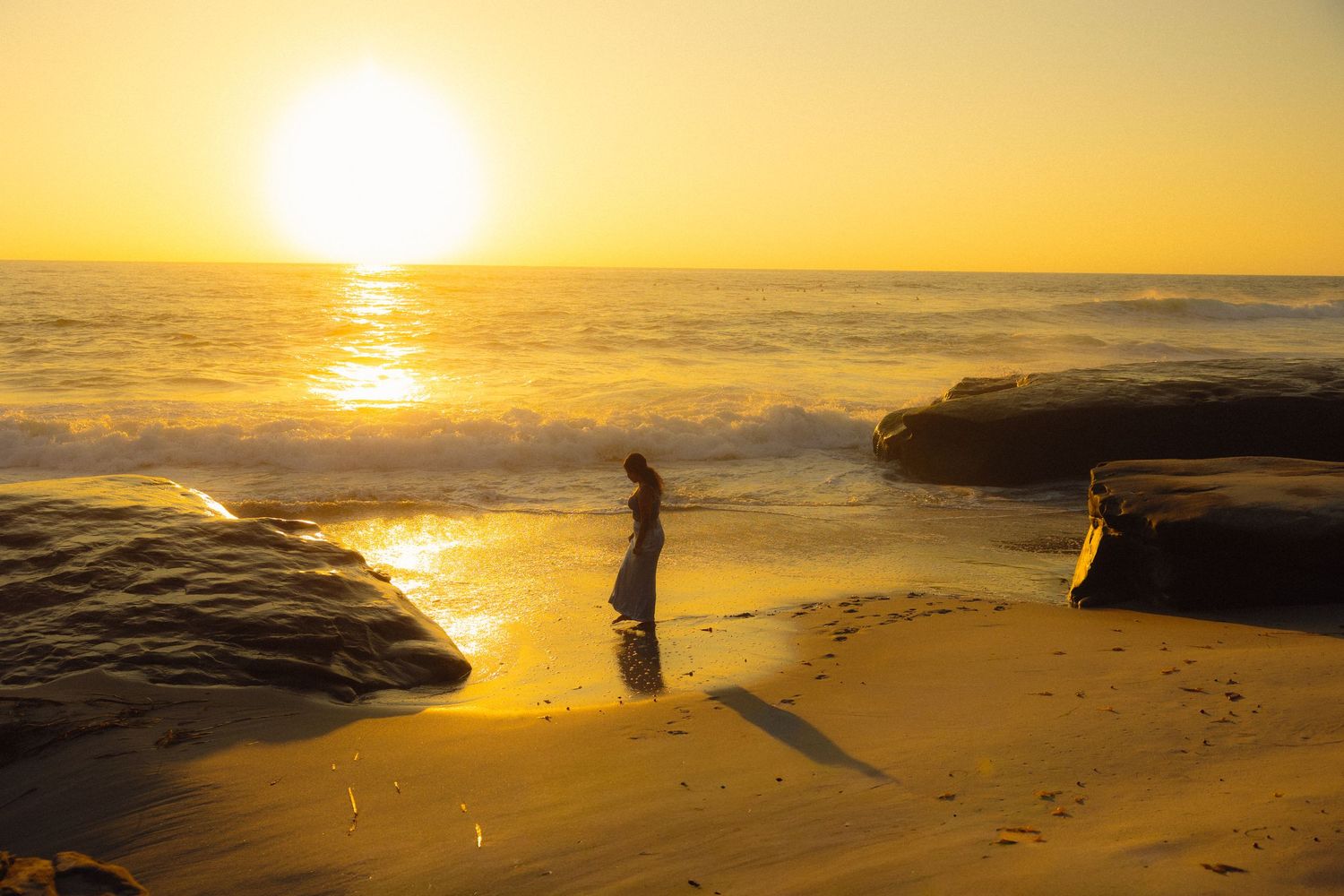 A golden sunset illuminates the beach as silhouettes walk along the shore with waves crashing against rocky cliffs.