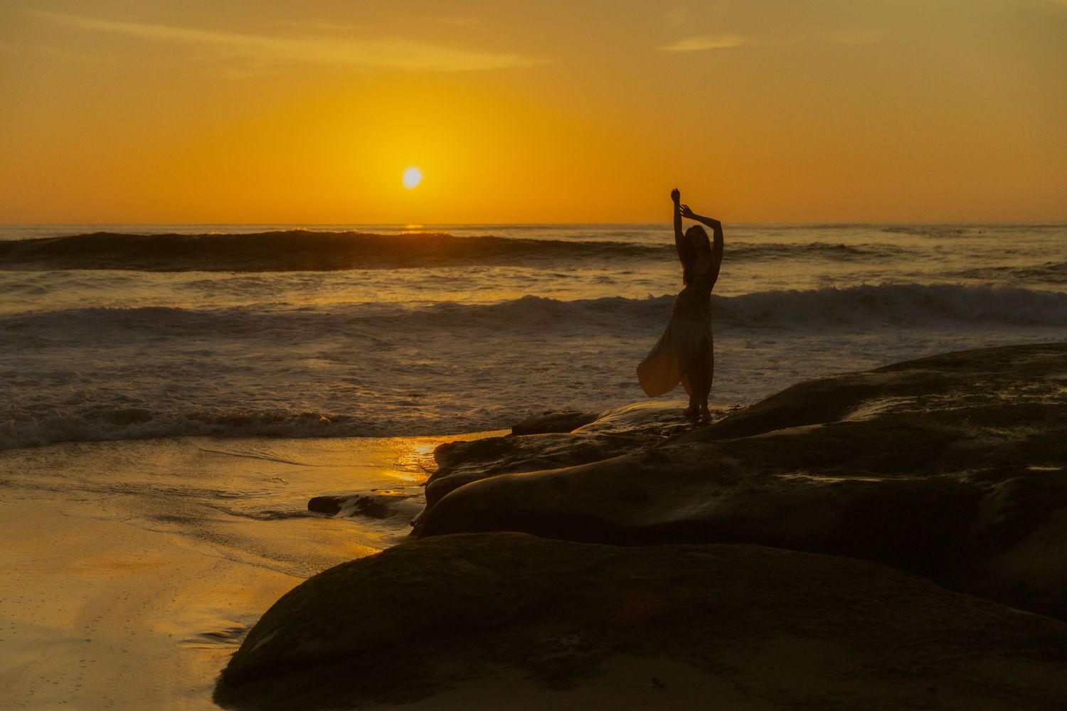A golden sunset illuminates the ocean horizon as waves crash against rocky coastal cliffs.