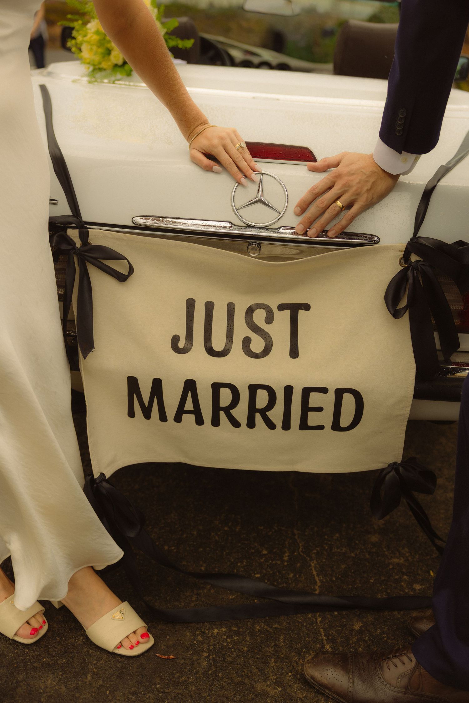 Newlyweds touch a Mercedes hood ornament above a Just Married sign at their wedding car.