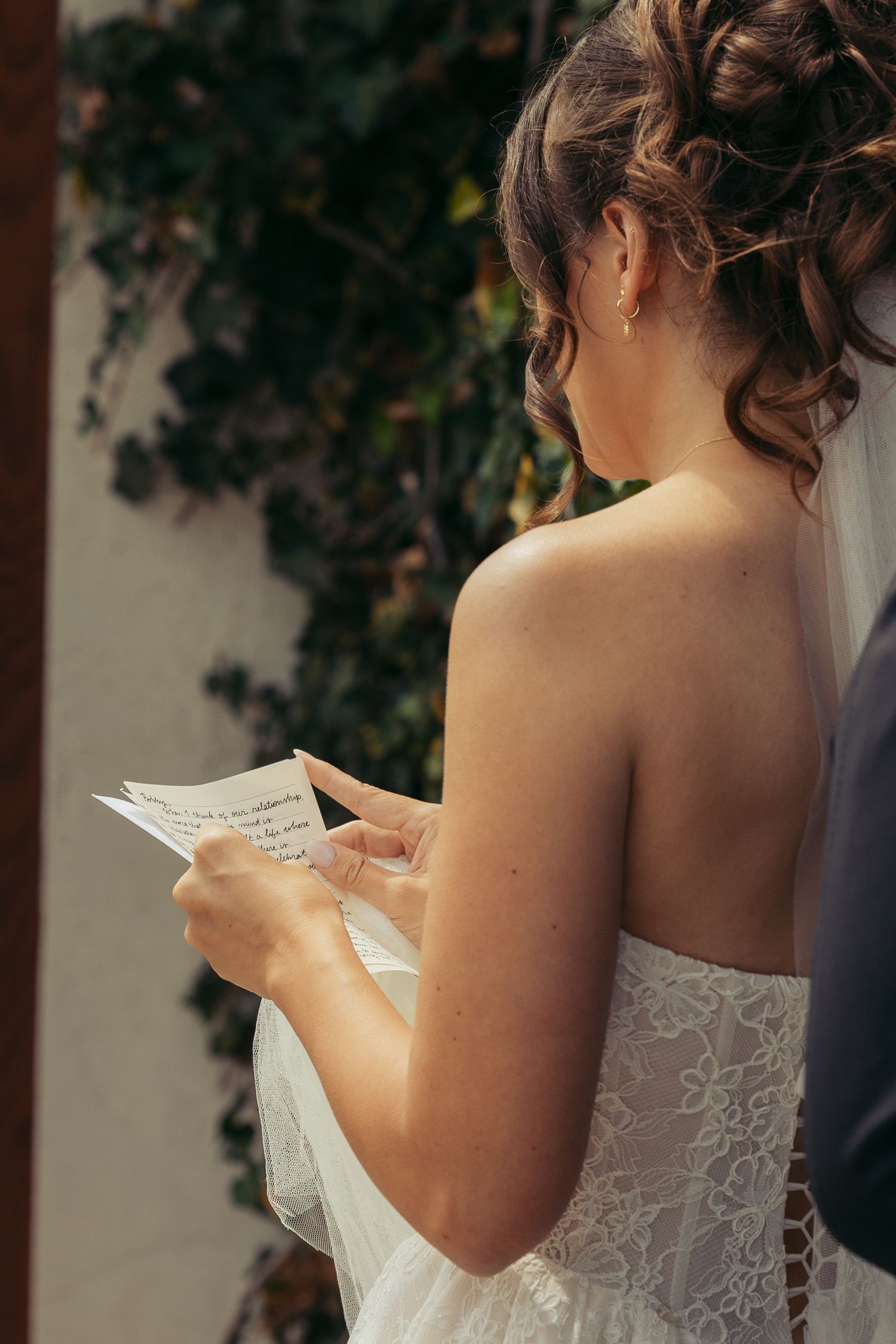 Bride in lace wedding dress reading handwritten vows, elegant updo hairstyle with veil, outdoor ceremony backdrop.