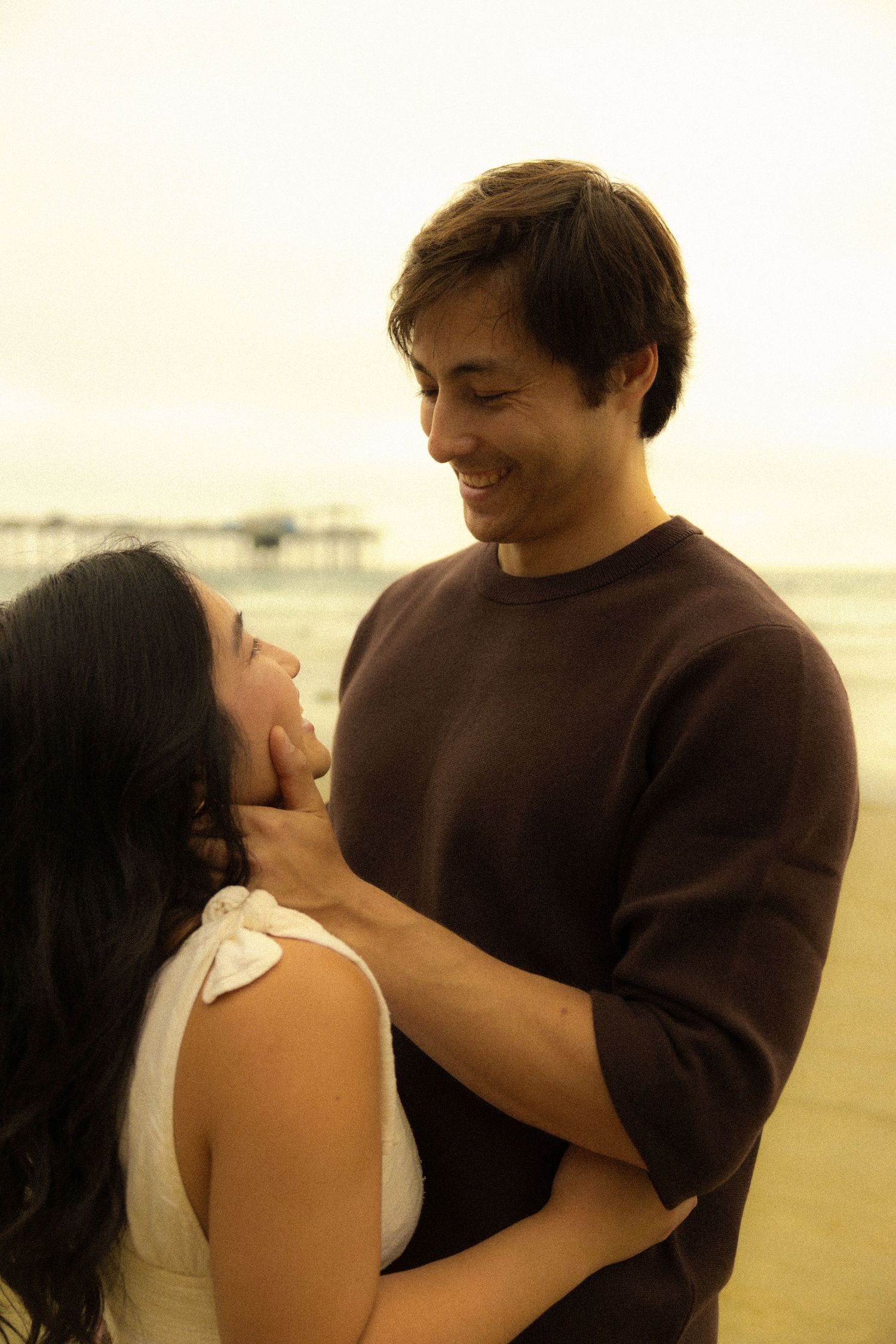 A romantic couple embraces on a misty beach at sunset, dressed in brown sweater and white top.