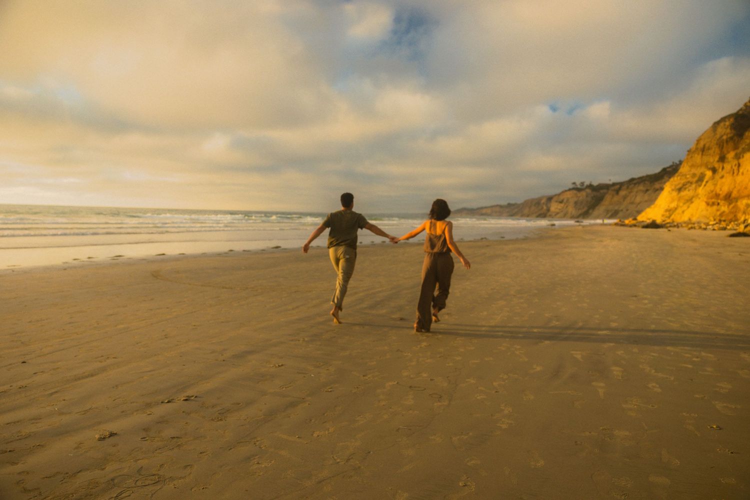 Two silhouetted figures walk along a sandy beach at sunset with golden cliffs in the background.