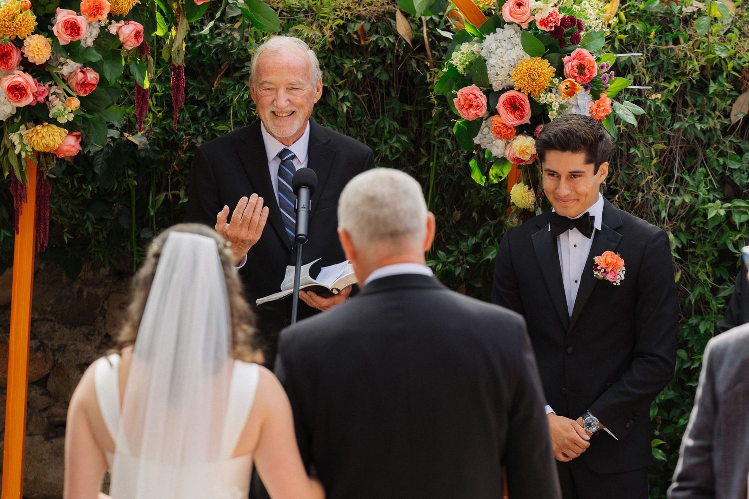 Outdoor wedding ceremony with groom in tuxedo, officiant smiling, and colorful floral arch backdrop.