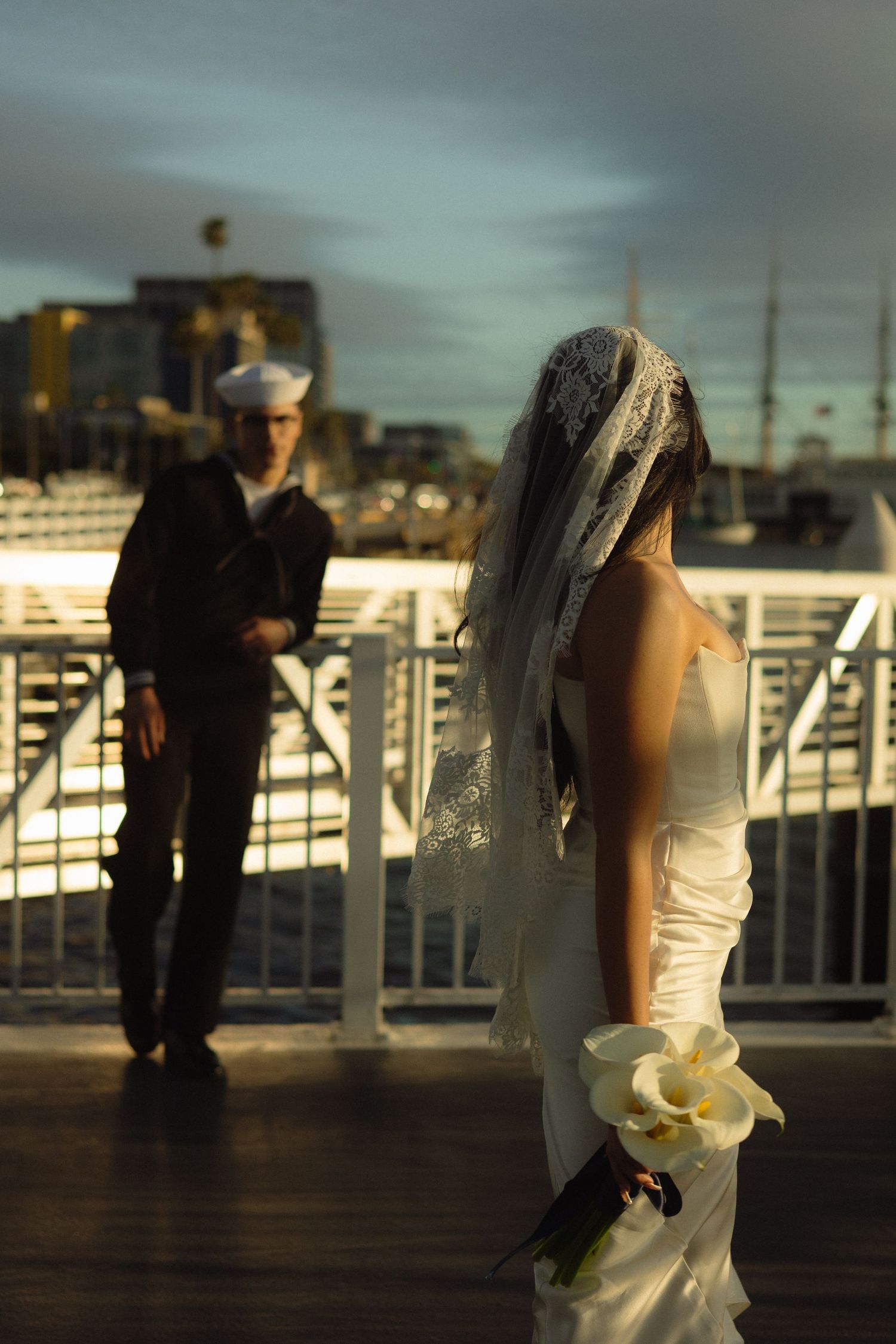 A bride and sailor stand on a deck at dusk with city lights in the background.