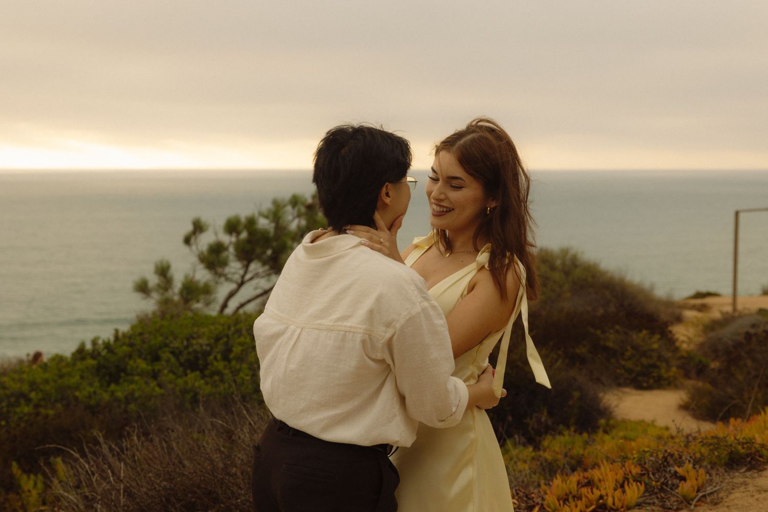 Romantic couple in white embraces during sunset photoshoot along scenic coastal cliffs.