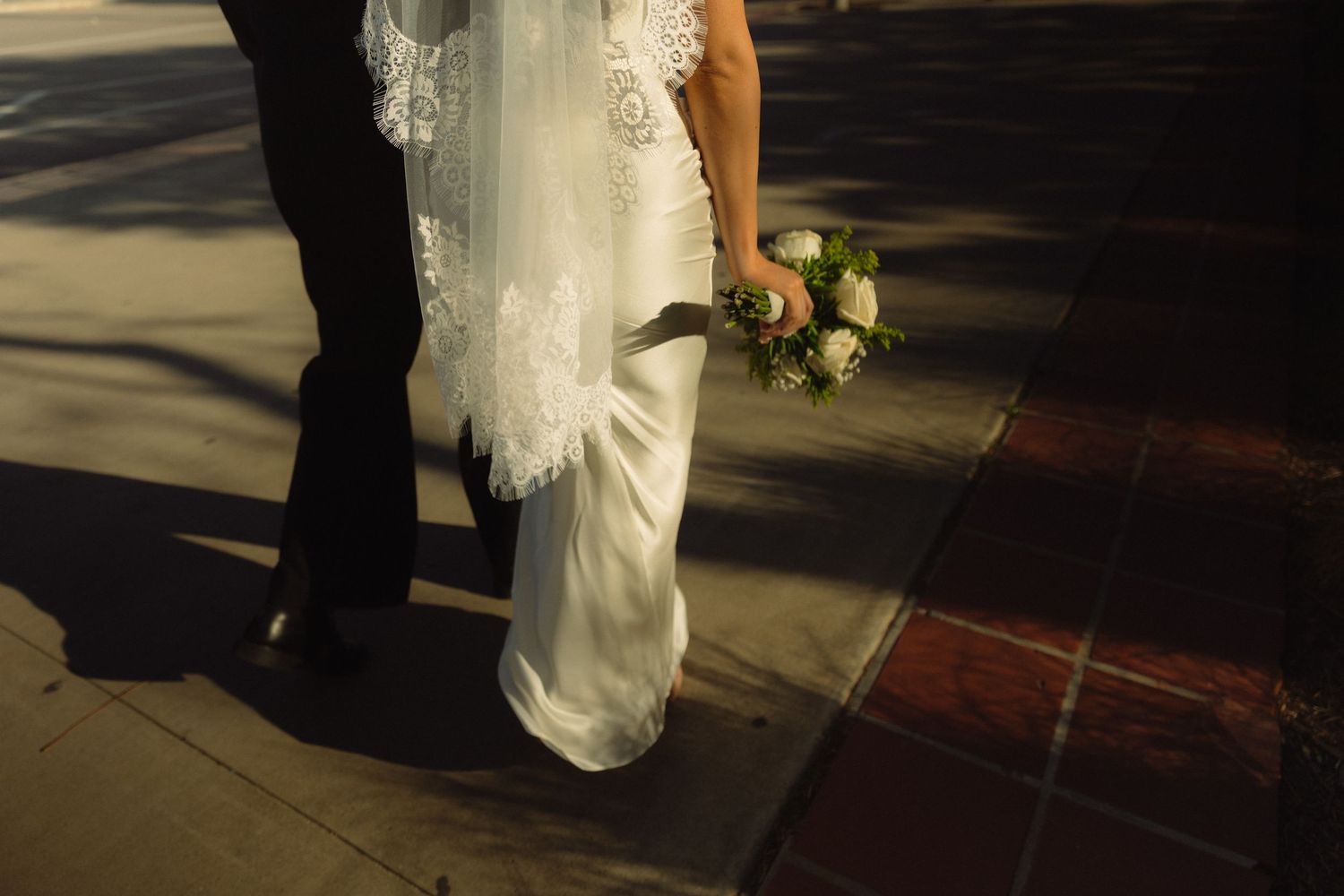 Cropped view of bride in white lace dress holding bridal bouquet while standing on brick sidewalk.