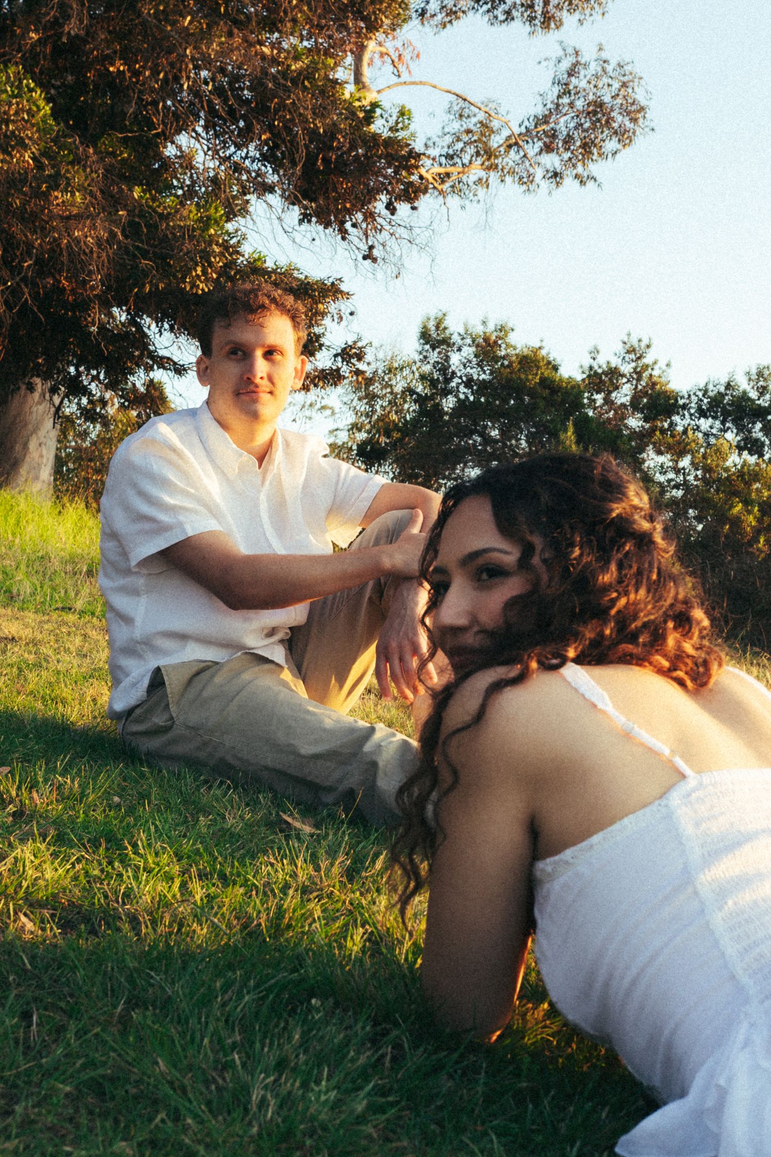 A couple sitting on green grass under a tree at golden hour, woman in white dress leaning forward, man in white shirt behind her.