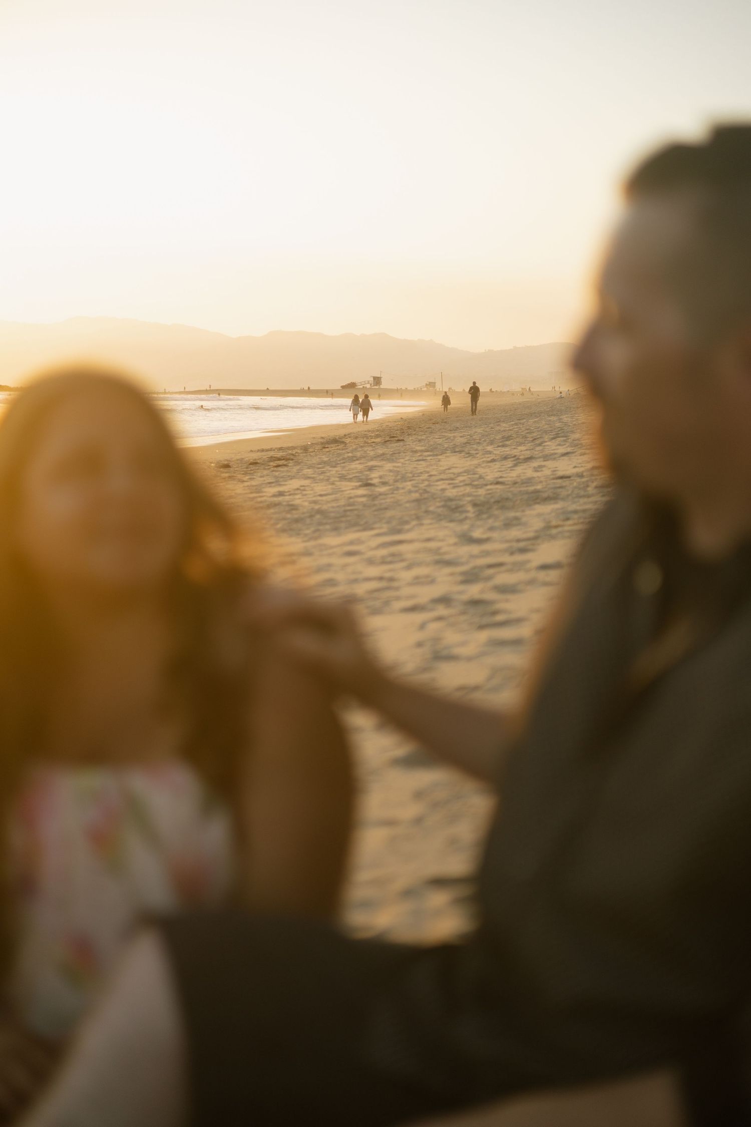 Couple on beach at sunset with distant figures walking along the shoreline in golden light.