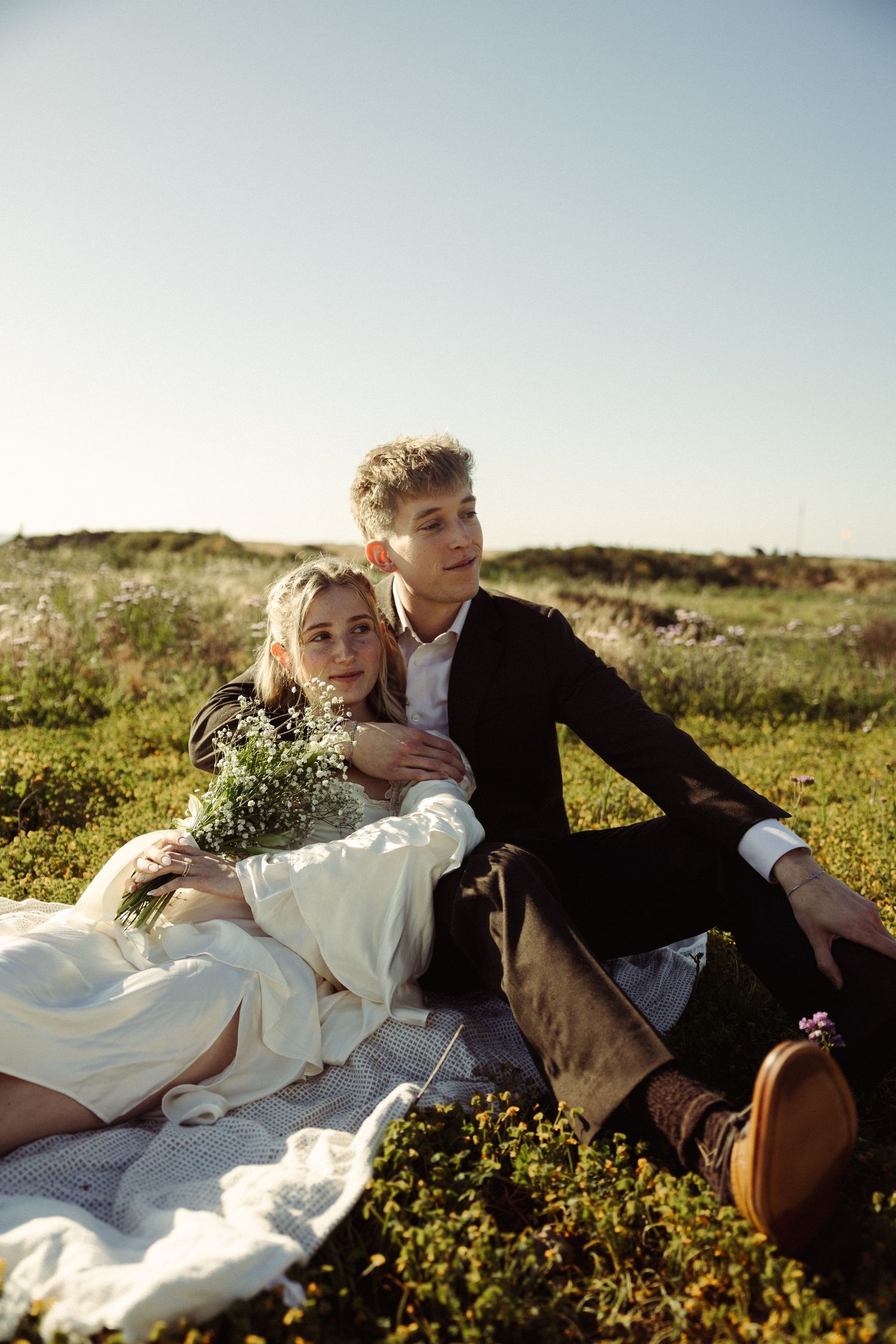 A young couple in wedding attire sits together on a blanket in a green field, the bride holding a wildflower bouquet.