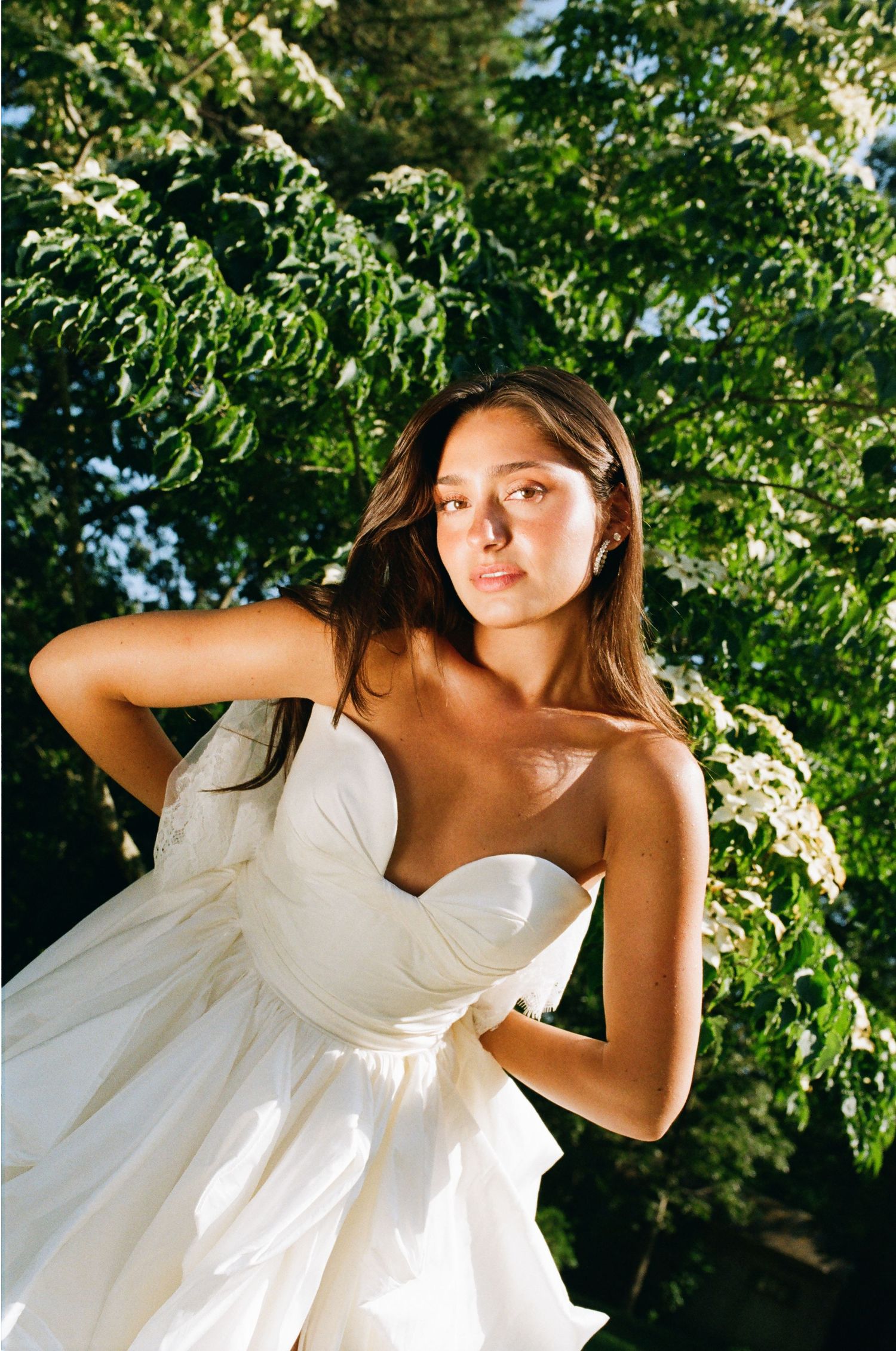 Young woman in a strapless white bridal gown poses outdoors among lush green trees in warm sunlight.