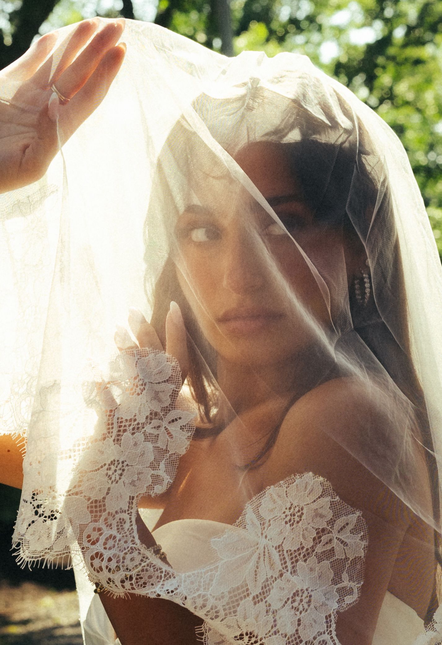 Bride gazes through lace-trimmed veil in golden sunlight with hand raised near face.