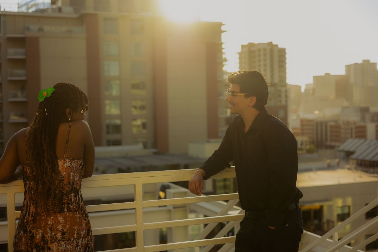Two people chat on a rooftop balcony at sunset with urban buildings in the background.