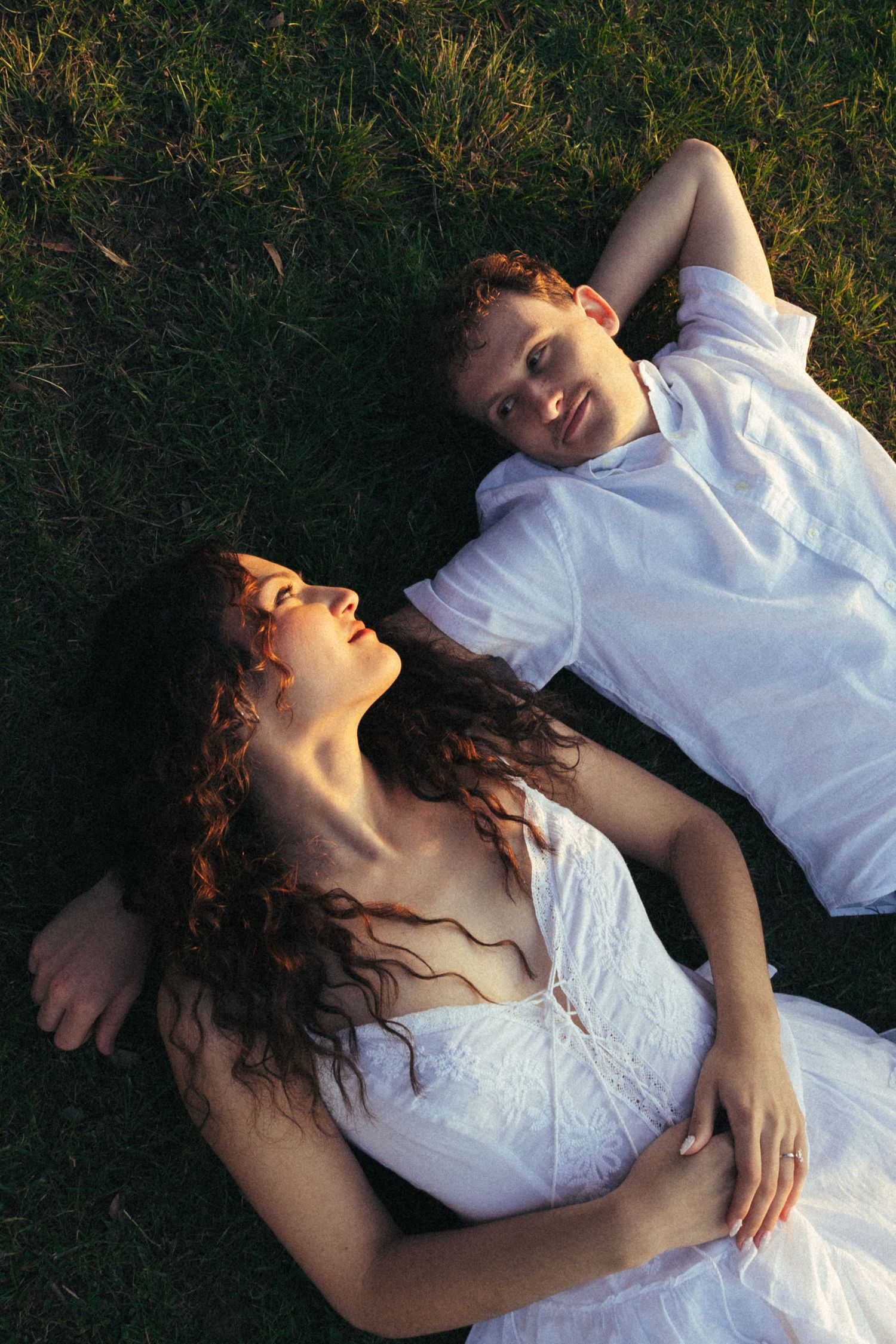 Couple in white outfits lying on green grass, woman with curly hair resting on man's chest in golden hour light.