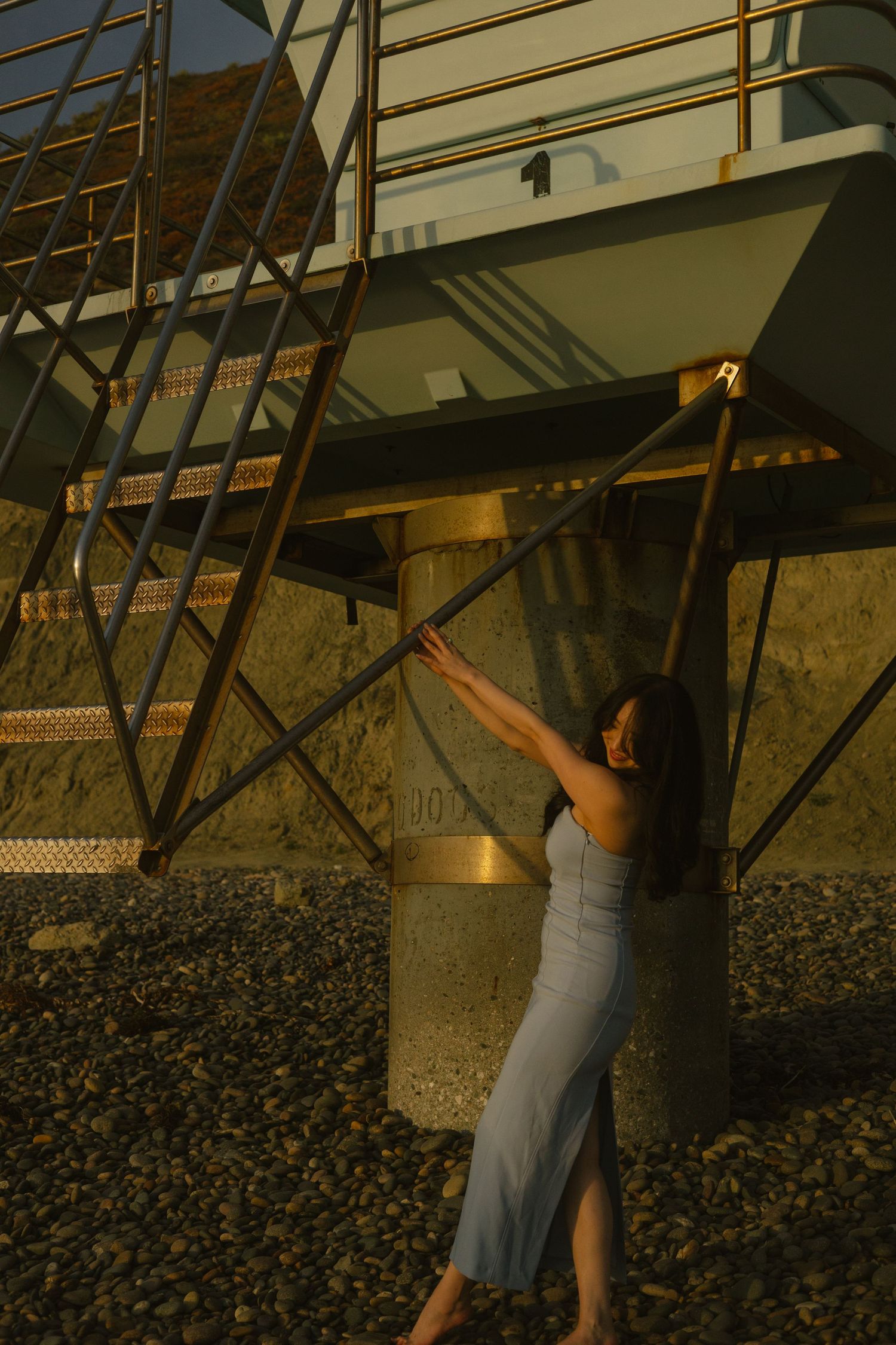 Person in light dress poses beneath industrial metal stairs during golden hour.