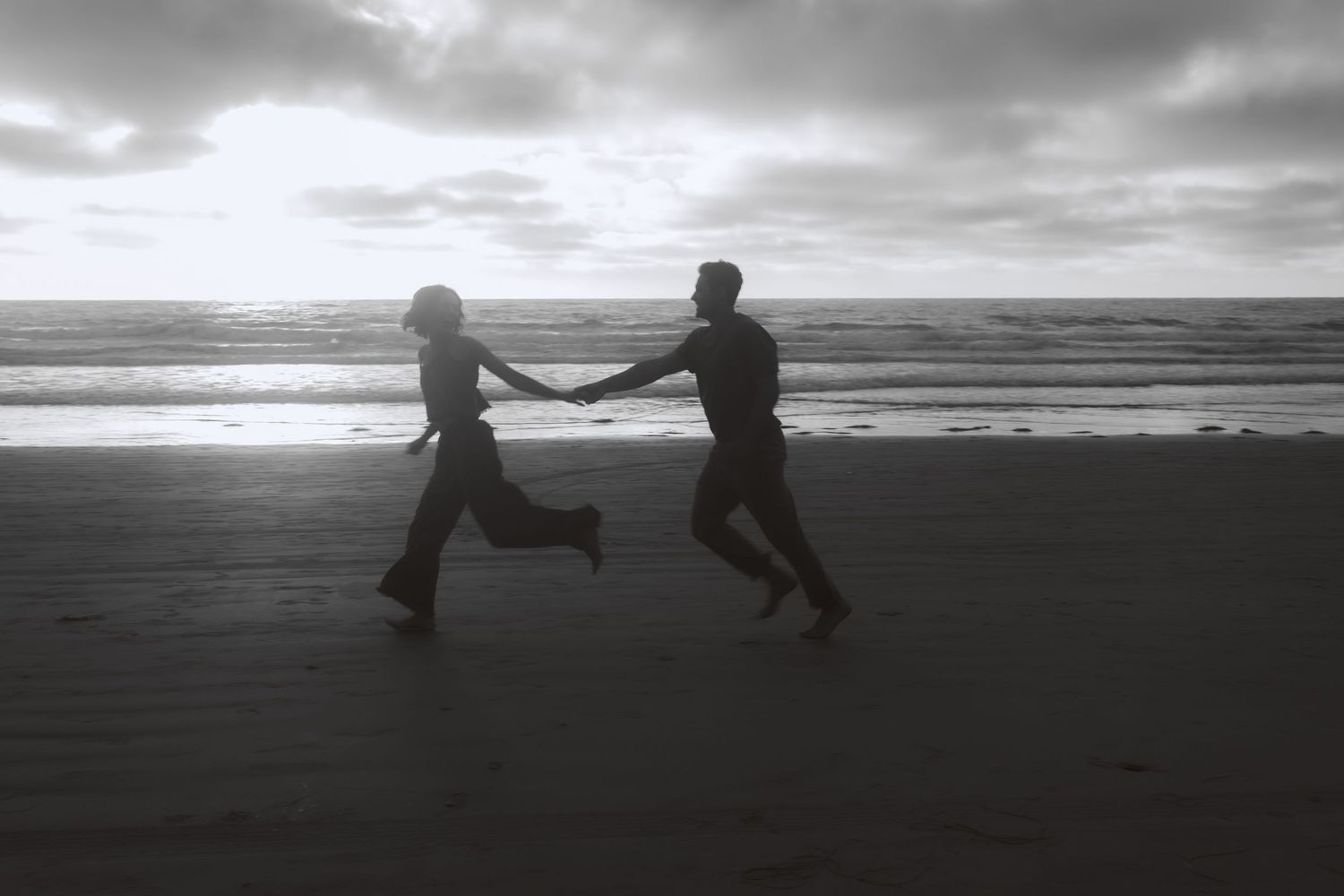 Silhouetted figures hold hands and dance along a misty beach at sunset creating a dreamy black and white scene.
