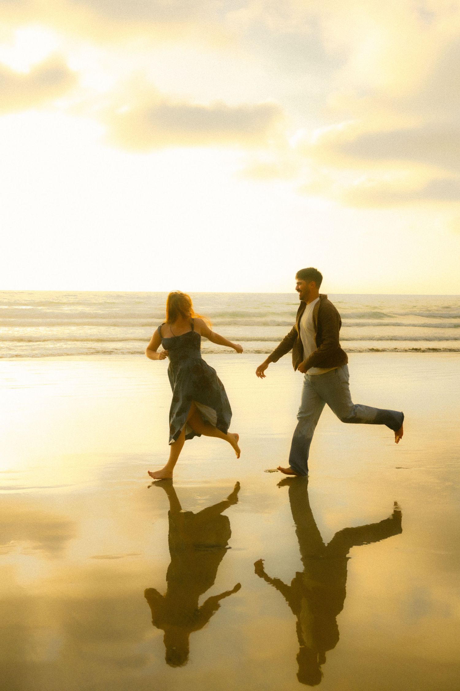 Silhouetted couple runs playfully along beach at sunset with their reflections mirrored in wet sand.