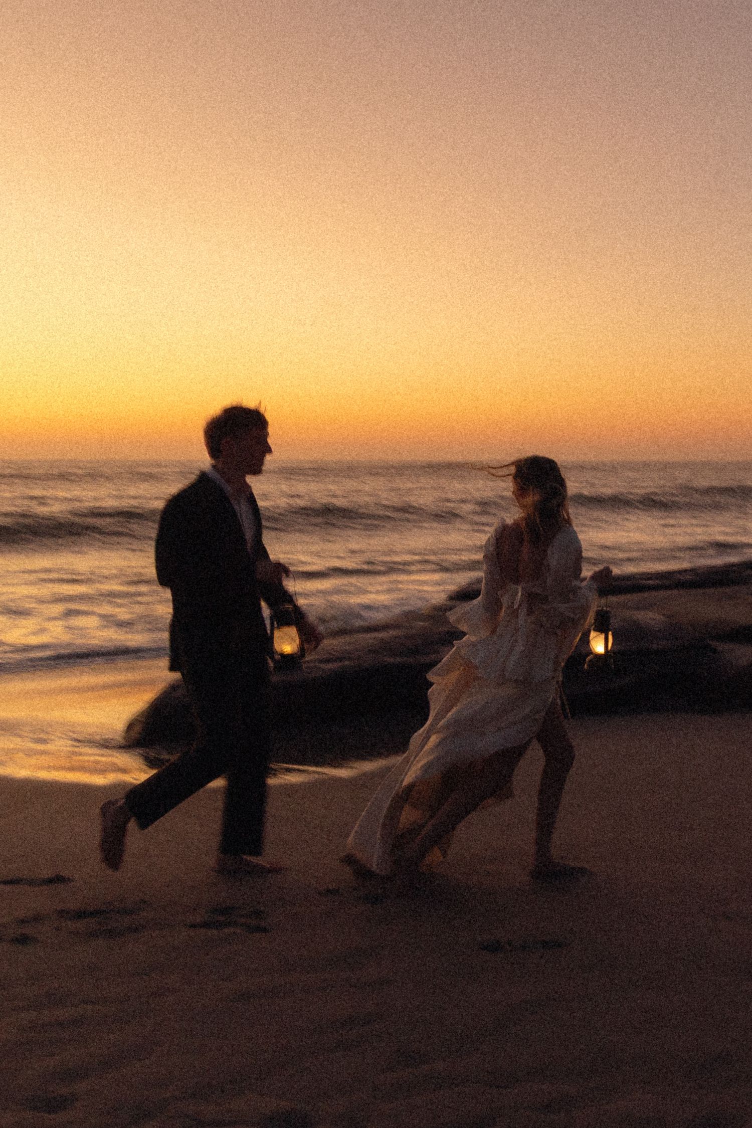 Couple in wedding attire running barefoot on a beach at sunset, holding lanterns with golden ocean horizon behind them.