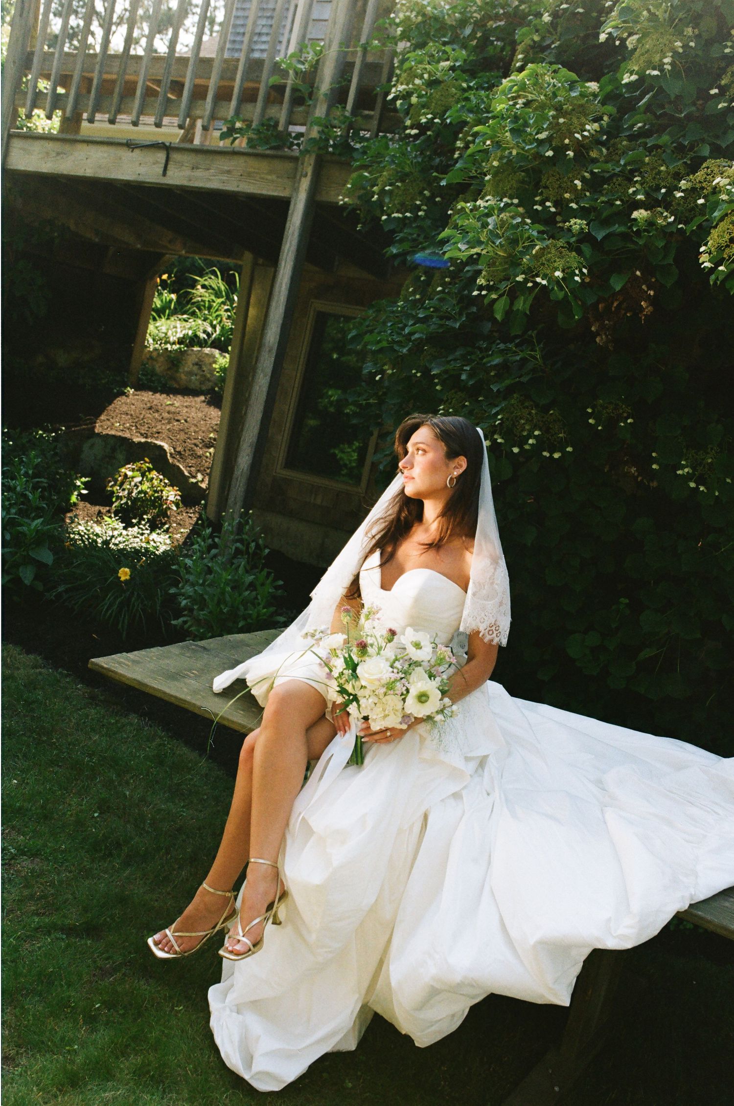 A bride in a white gown and veil sits outdoors holding a bouquet, gazing upward in a lush garden setting.