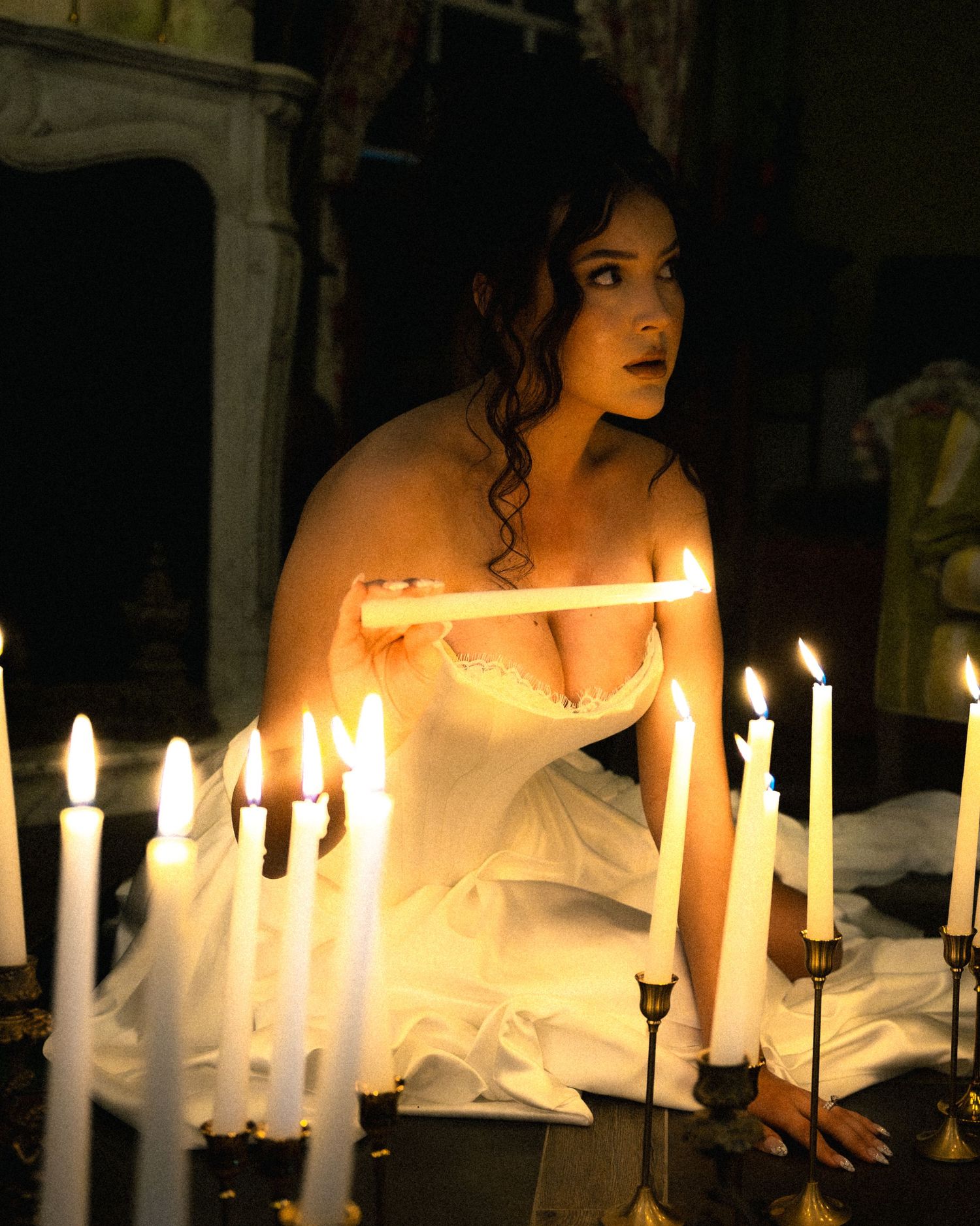 Figure in white dress illuminated by multiple candlesticks creating an ethereal ambiance in dark setting.