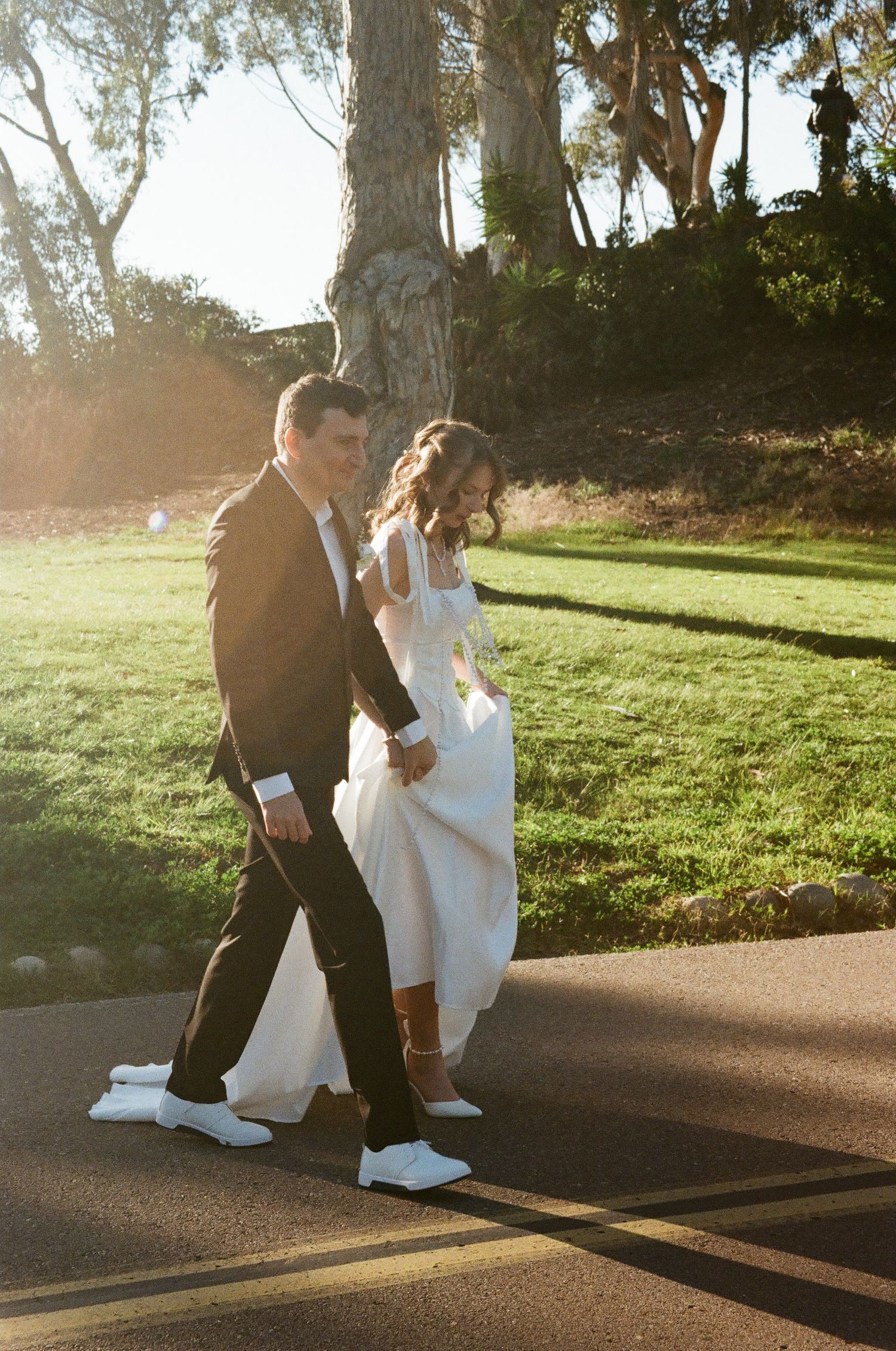 A couple in formal attire dances together on a sunlit path in a park during golden hour.